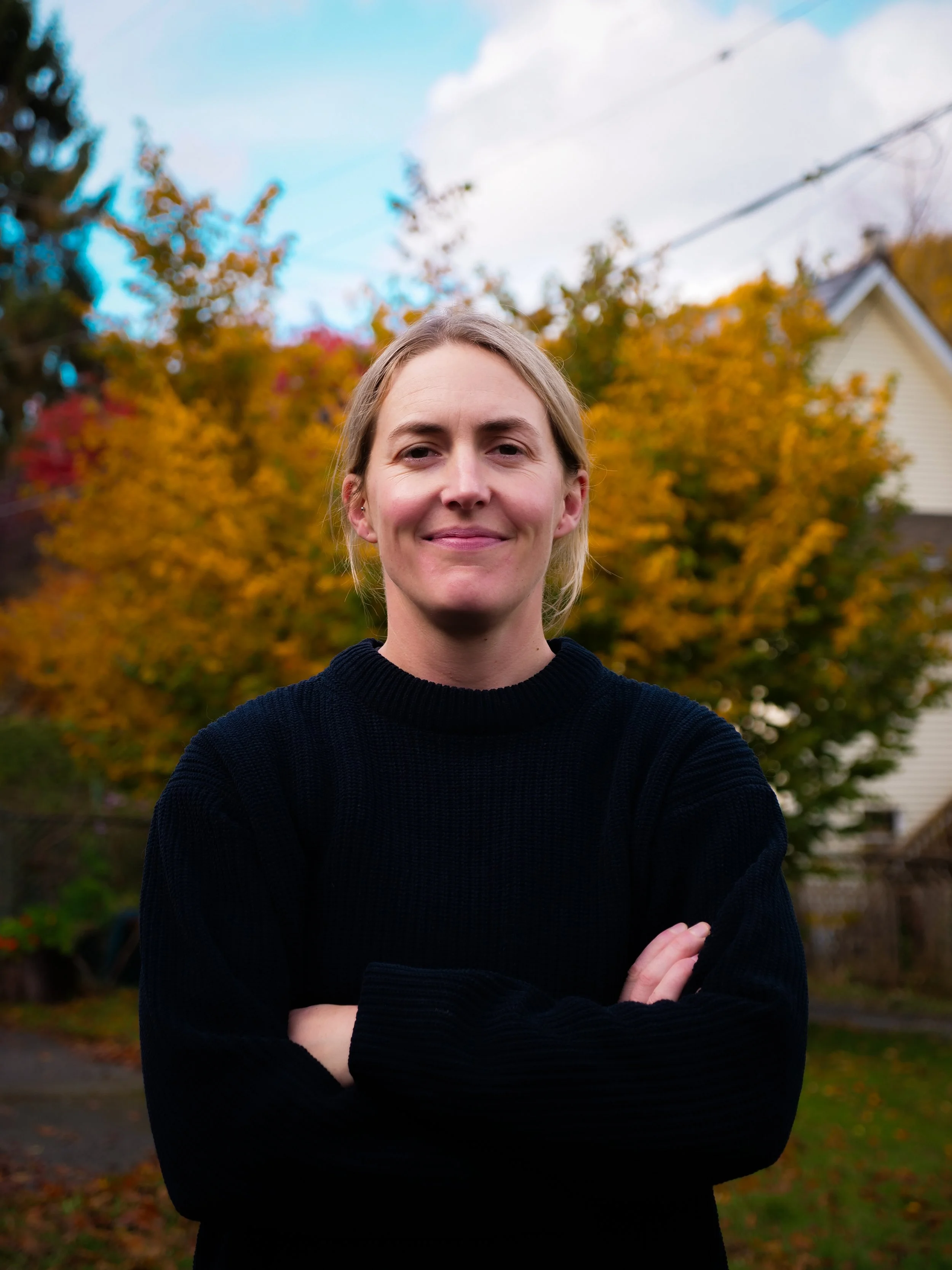 A woman standing outdoors with autumn trees in the background, smiling with arms crossed, wearing a black sweater.
