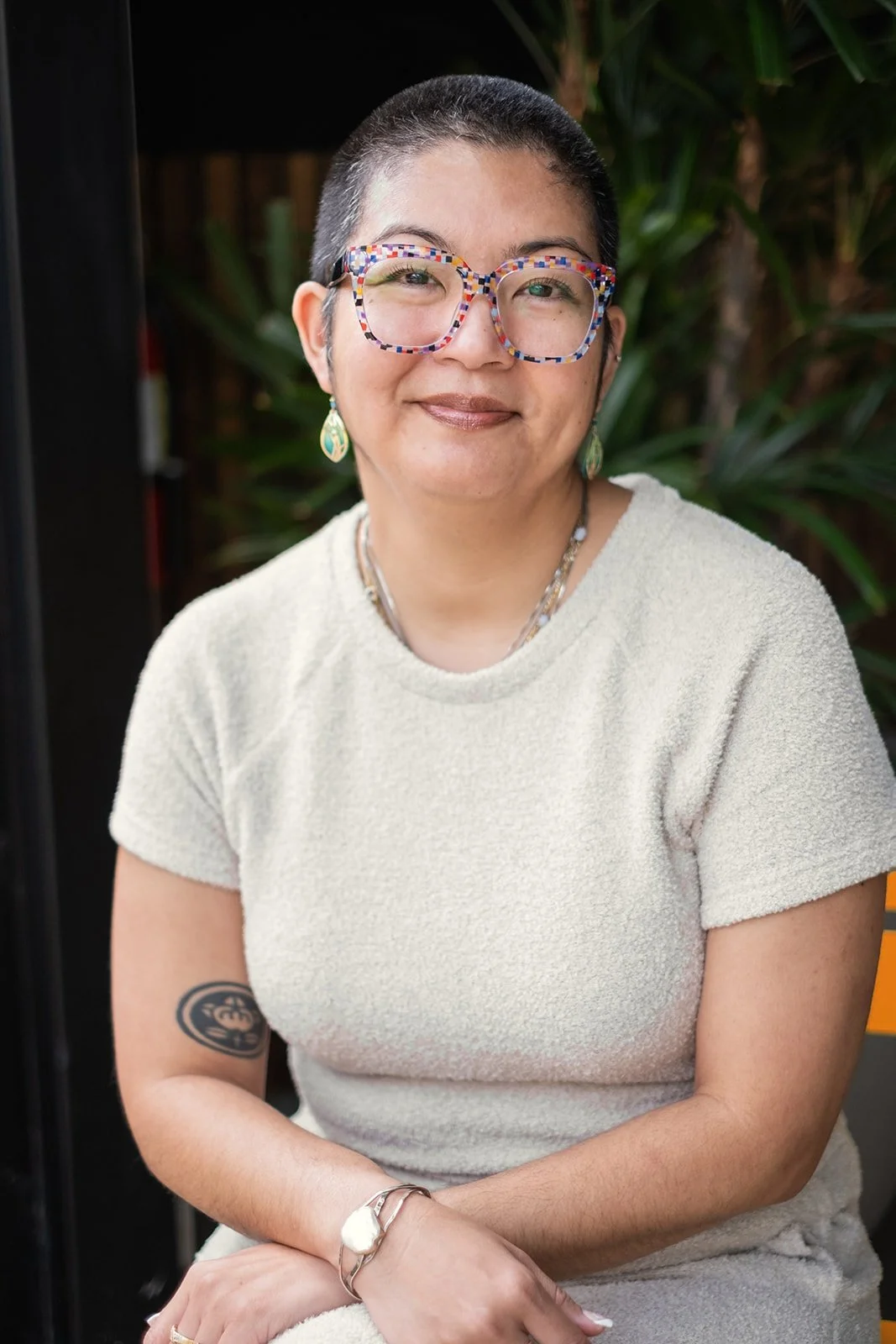 Headshot of Kylie with short hair and glasses sitting outside, smiling at the camera. She wears a light-colored textured top, earrings, a necklace, and a bracelet, with a tattoo on her right arm and green foliage in the background.