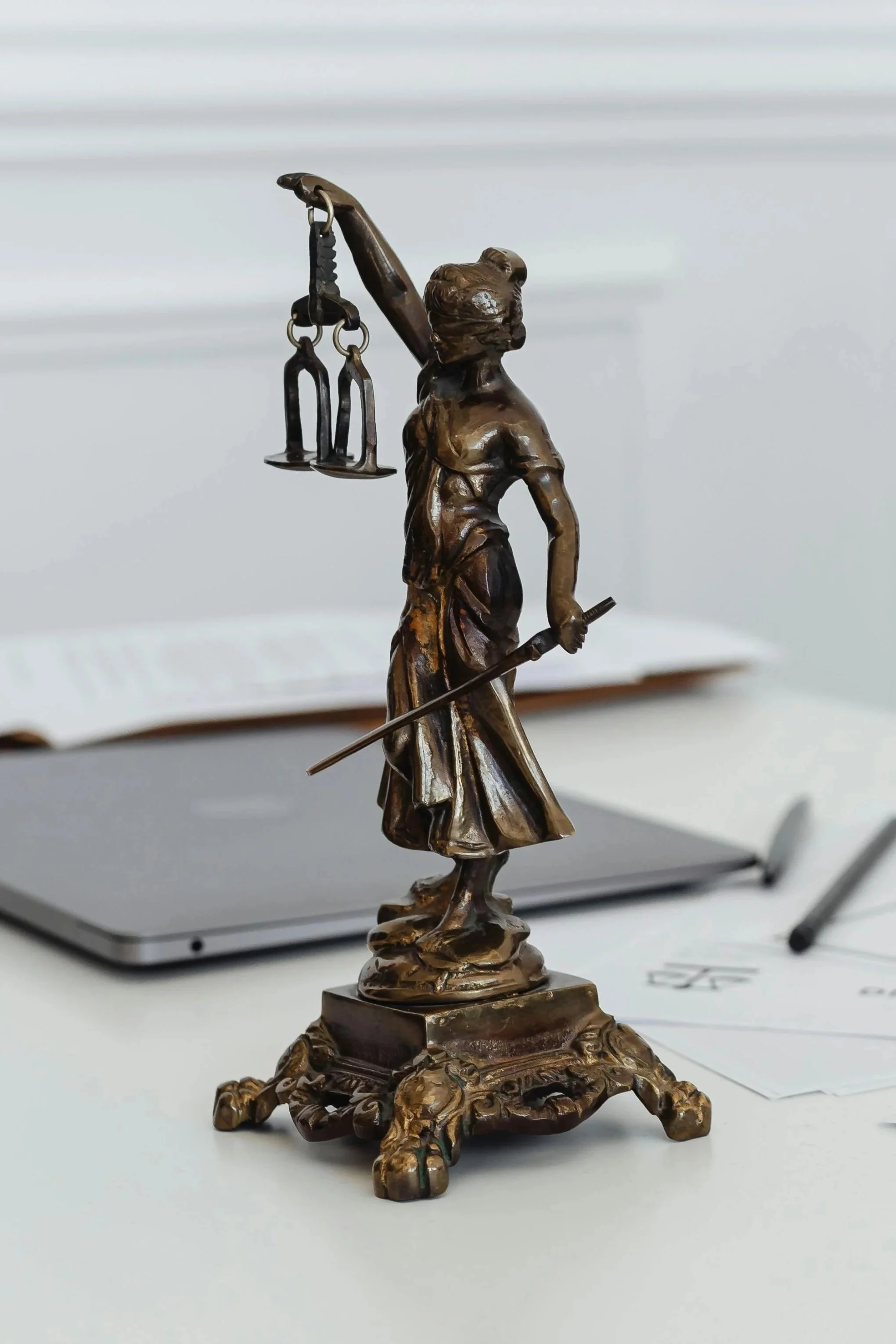 Bronze statue of Lady Justice holding scales and a sword, placed on a desk with a laptop and papers in the background.