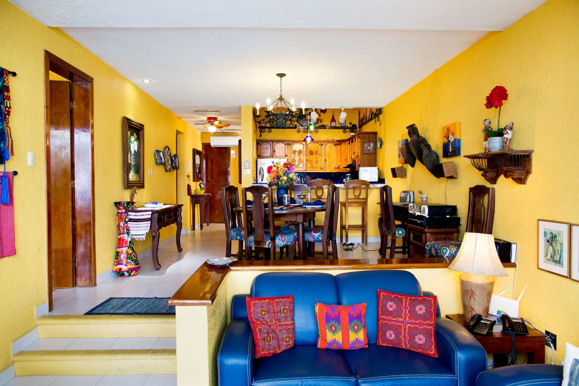 Cozumel vacation rental living room with yellow walls, blue sofa with colorful pillows, dining table with chairs, kitchen in background, and decorative items on the walls.
