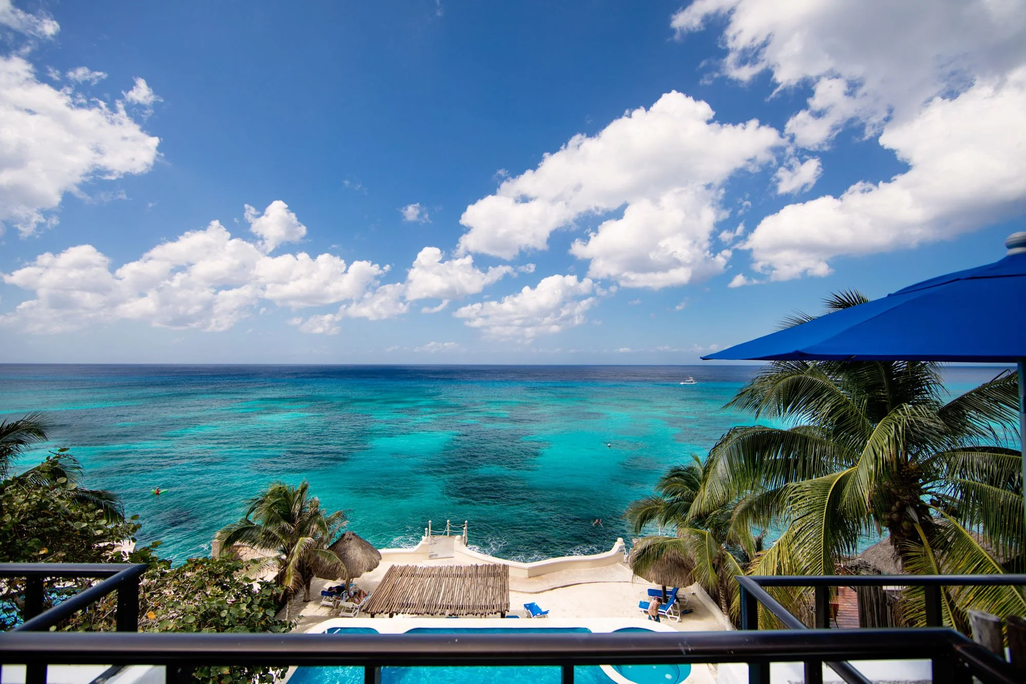 View from a Cozumel vacation rental balcony over a tropical beach with blue ocean waters, palm trees, blue umbrellas, and thatched huts under a partly cloudy sky.