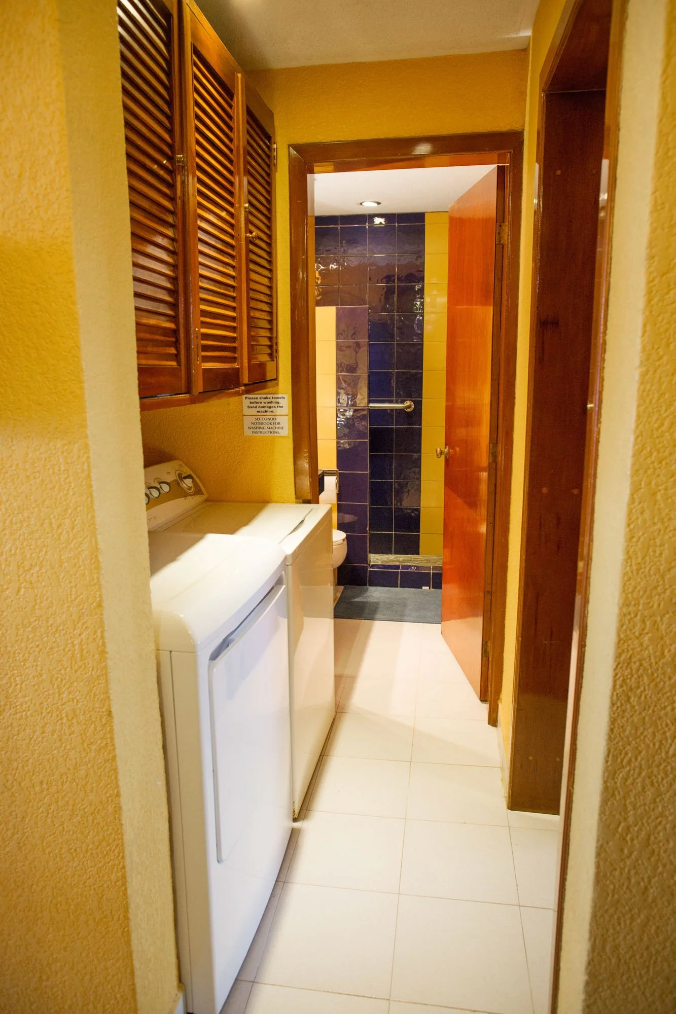 View of a laundry room with a washer and dryer, a wooden cabinet, and a doorway leading to a bathroom with dark blue and yellow tiled walls.