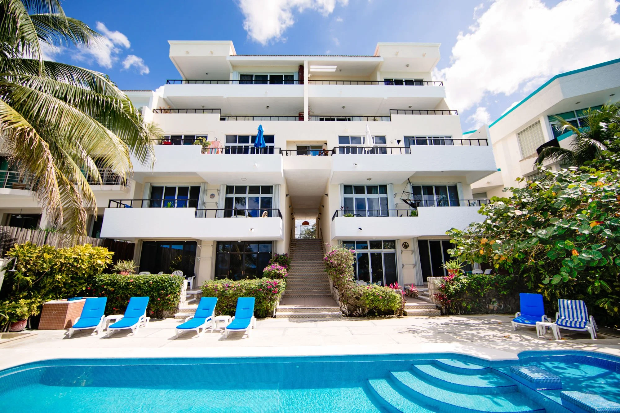 View of a Cozumel vacation rental with multiple balconies, a swimming pool in the foreground, and lush tropical plants and blue sky.