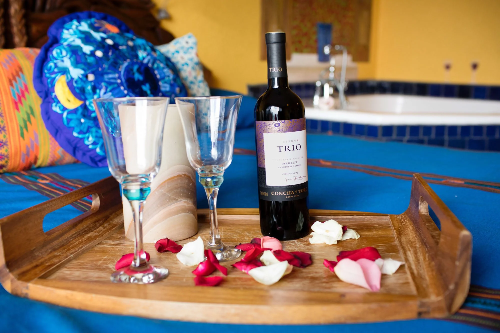 A wooden tray on a blue tablecloth holds a bottle of red wine, two empty wine glasses, and scattered rose petals. In the background, colorful cushions and a bathtub are visible, suggesting a cozy, romantic setting.