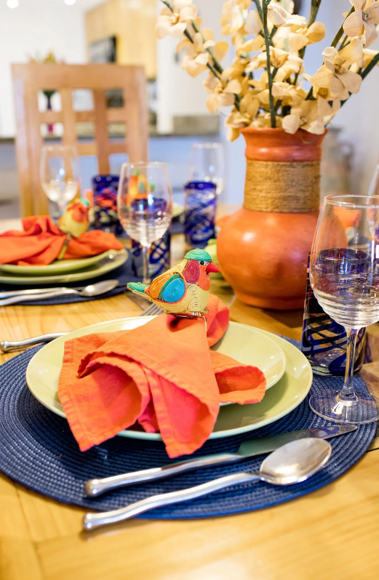 A festive dining table with orange napkins, colorful napkin rings shaped like turkeys, wine glasses, a large earthy vase with dried cream-colored flowers, and a small ceramic bird figurine. The table is set on a blue placemat with wooden chairs in th