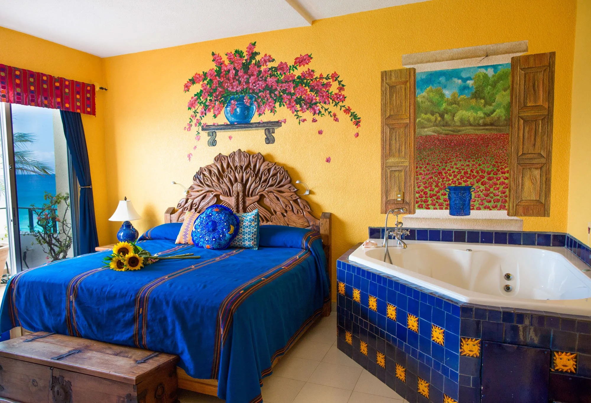 Bright yellow bedroom with a carved wooden bed, blue bedding, and sunflowers on the bed. Decorative wall paintings include a pink flower vase and a field of red flowers through a window. Blue-tiled jacuzzi next to the bed.