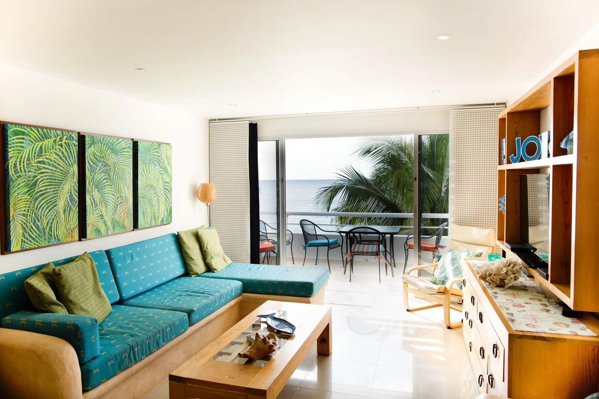 Living room with a large blue sofa, green pillows, and a painting of green hurricane leaves above. Sliding glass door leading to a balcony with a table and chairs, overlooking the ocean. Light-colored walls and tiled floor.