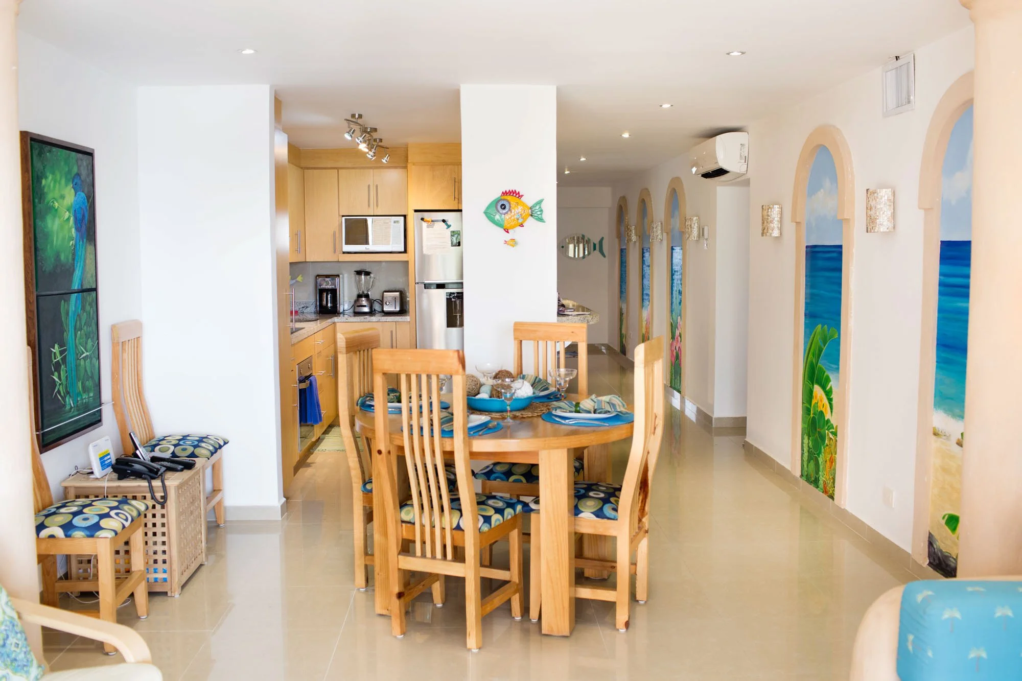 Dining area with wooden table and chairs set for a meal, in an open-plan kitchen and living space with tropical ocean-themed wall murals and beach decor.