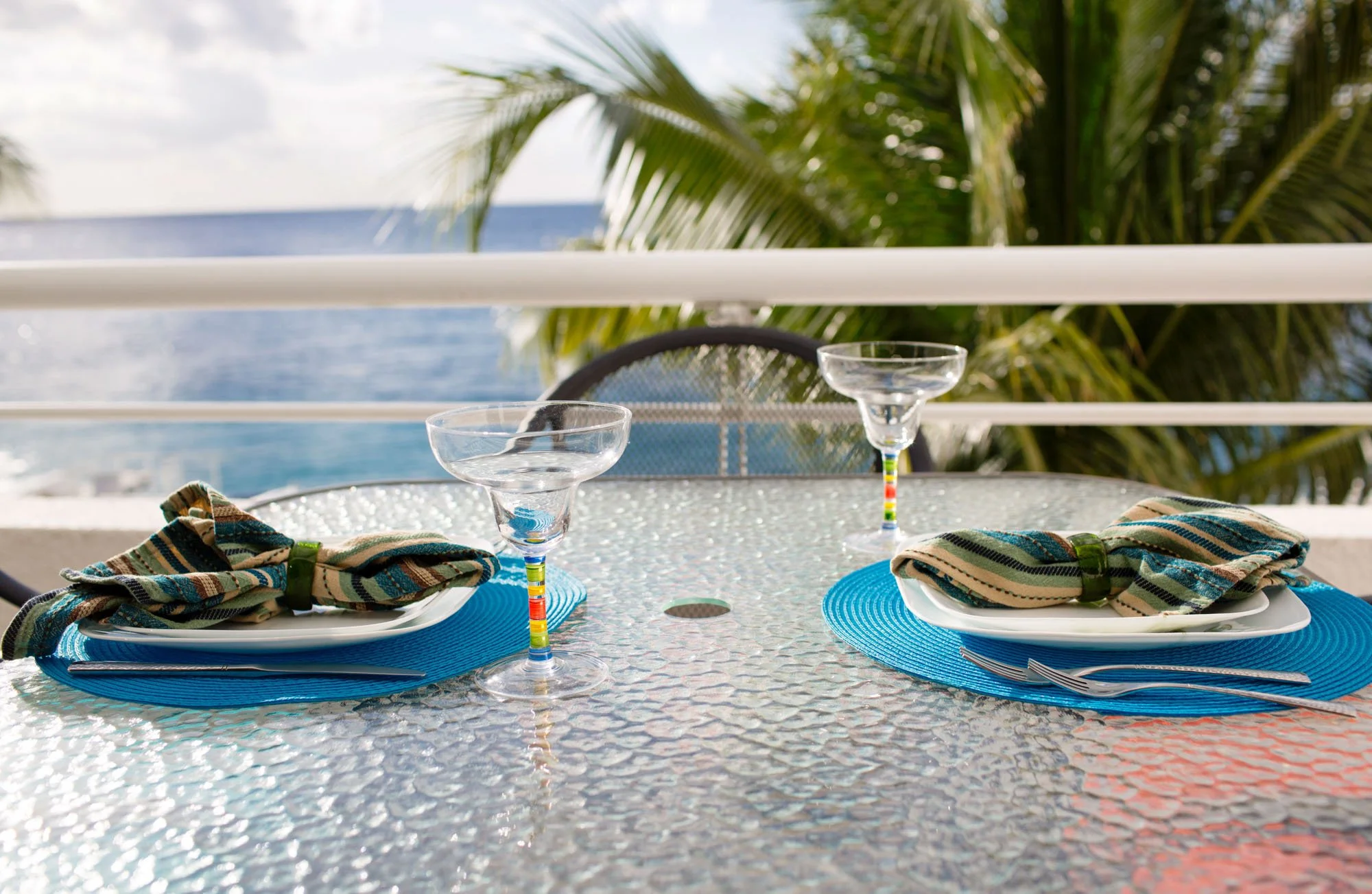 Table set outdoor with two place settings, glassware, and napkins, overlooking palm trees and the ocean.