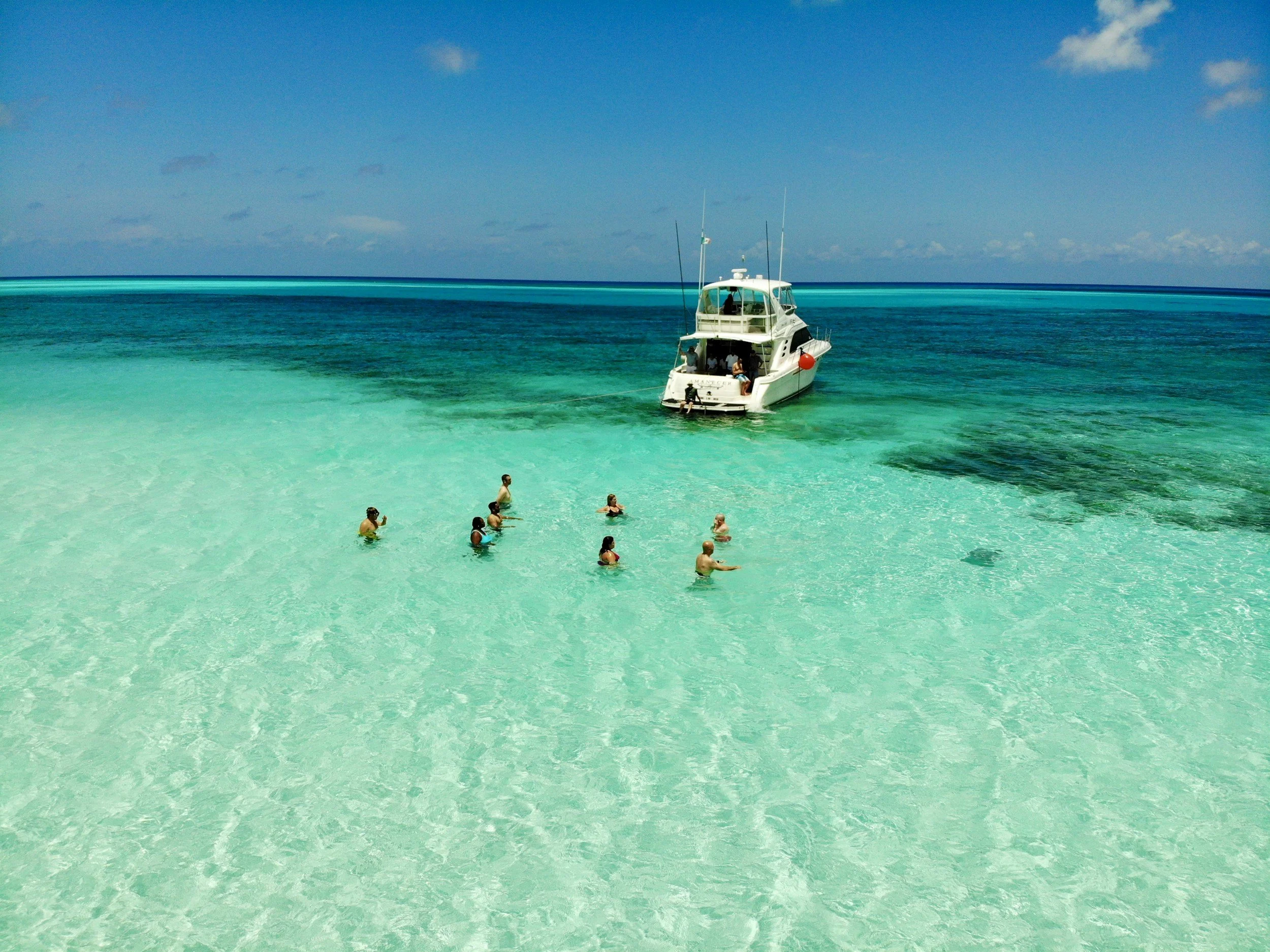 People swimming in clear turquoise water near a white boat in a tropical ocean under a partly cloudy sky.