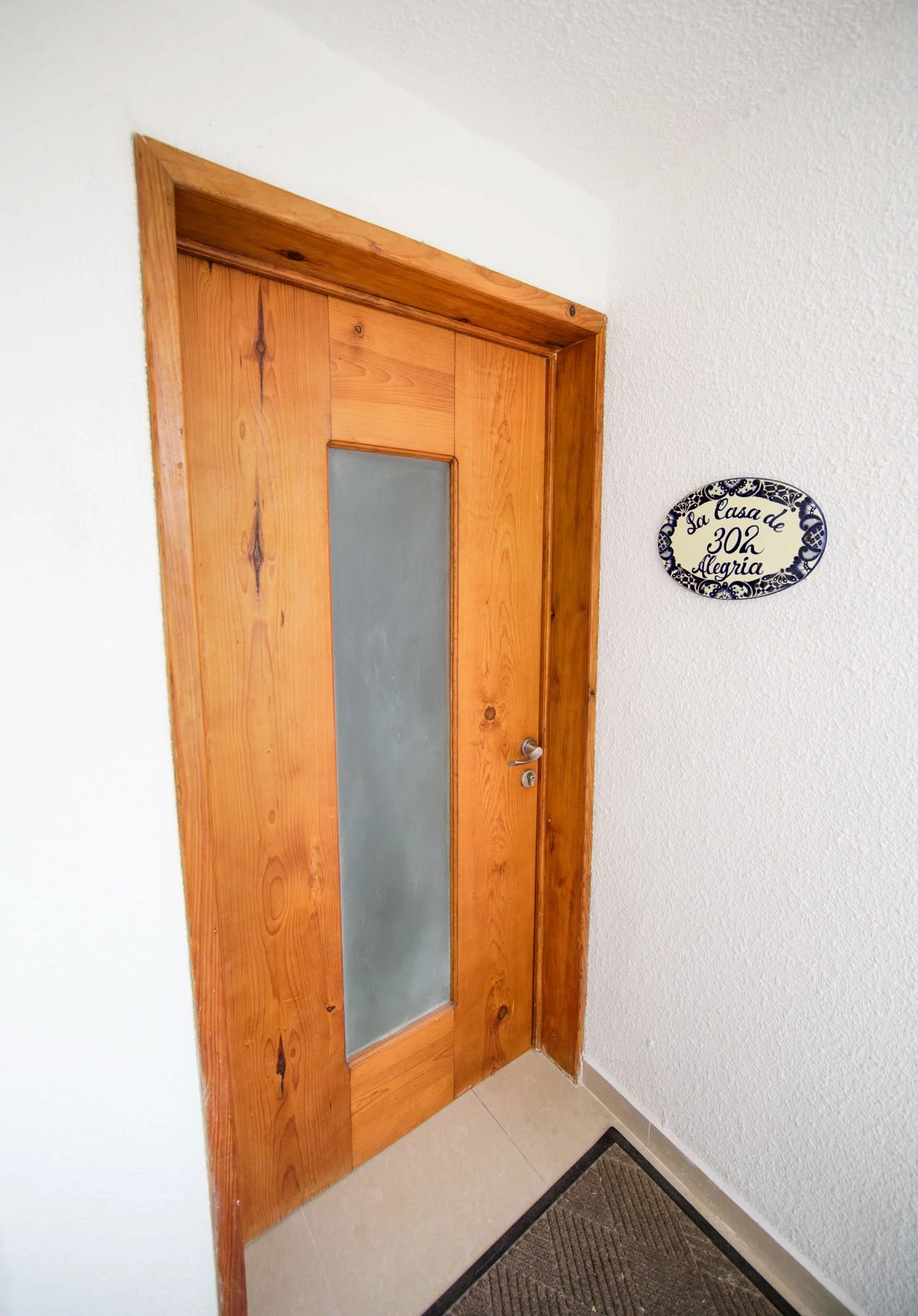 Wooden door with frosted glass panel in a white-walled entryway, and a decorative sign next to it that reads in Spanish: 'La casa de 302 alegría'.
