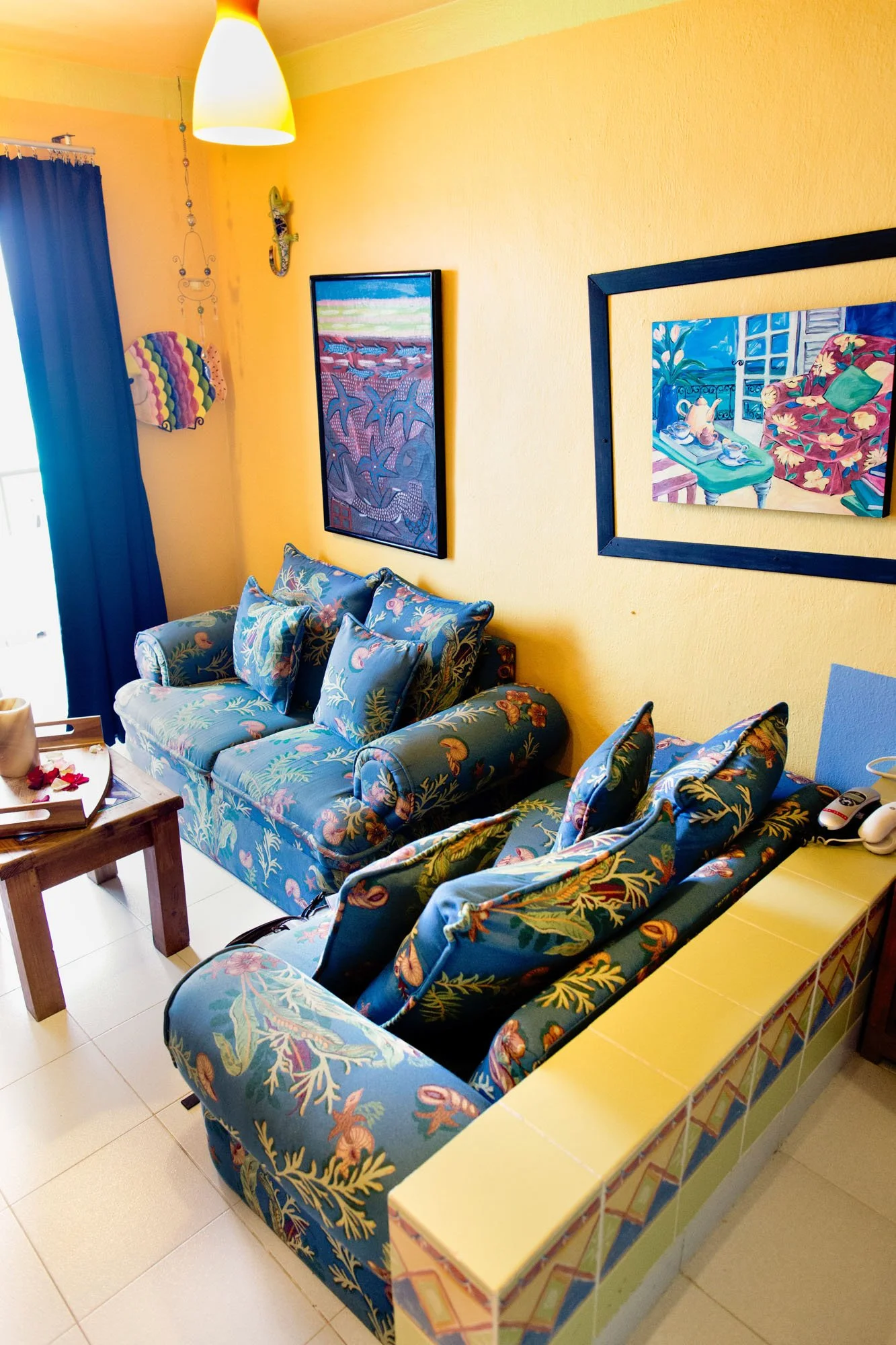 Living room with yellow walls, a floral-patterned sofa, and matching chairs, a wooden coffee table, and framed artwork on the walls. Blue curtains cover a window.