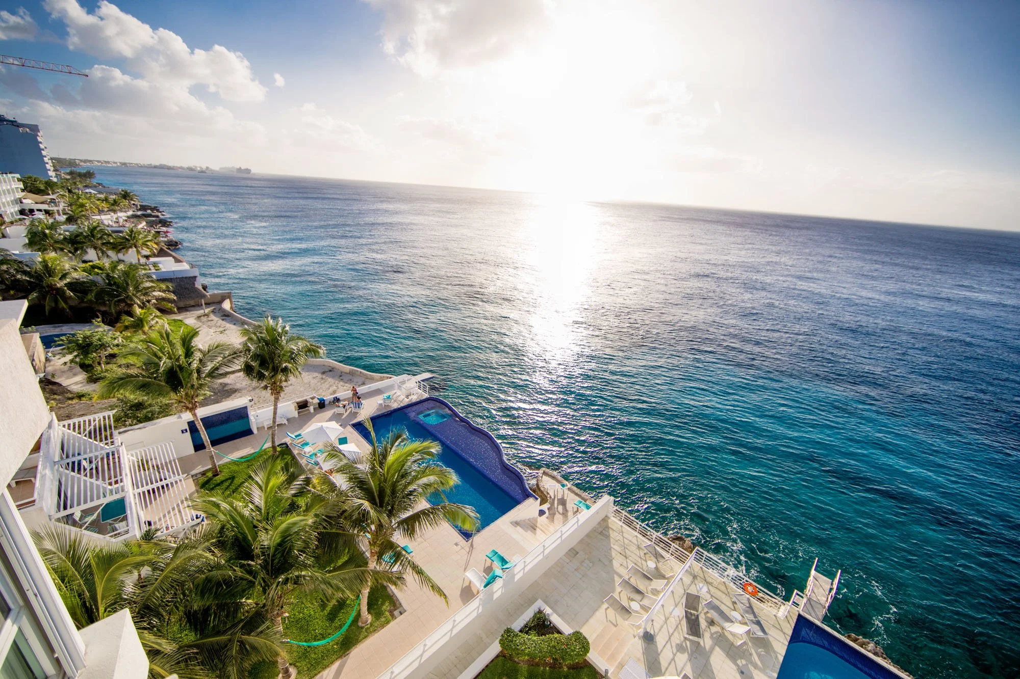 Aerial view of a luxurious seaside Cozumel vacation rental with a swimming pool, palm trees, and outdoor lounge chairs overlooking the ocean during sunset.