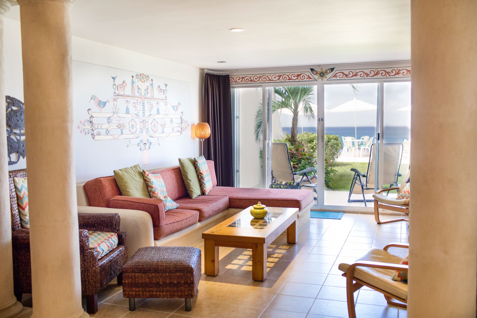 Living room with a pink sectional sofa, a wicker armchair, a wooden coffee table, and sliding glass doors leading to a balcony with outdoor chairs and an ocean view at a Cozumel vacation rental.