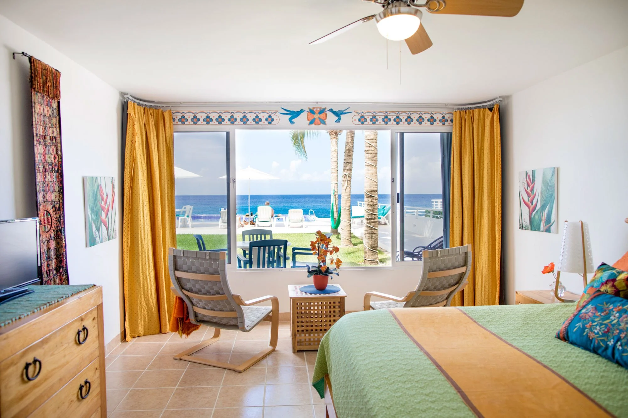 View from a hotel room looking out at the ocean with a balcony, chairs, play swing, and palm trees, featuring a large window, bed, and tropical decor inside at a Cozumel vacation rental.
