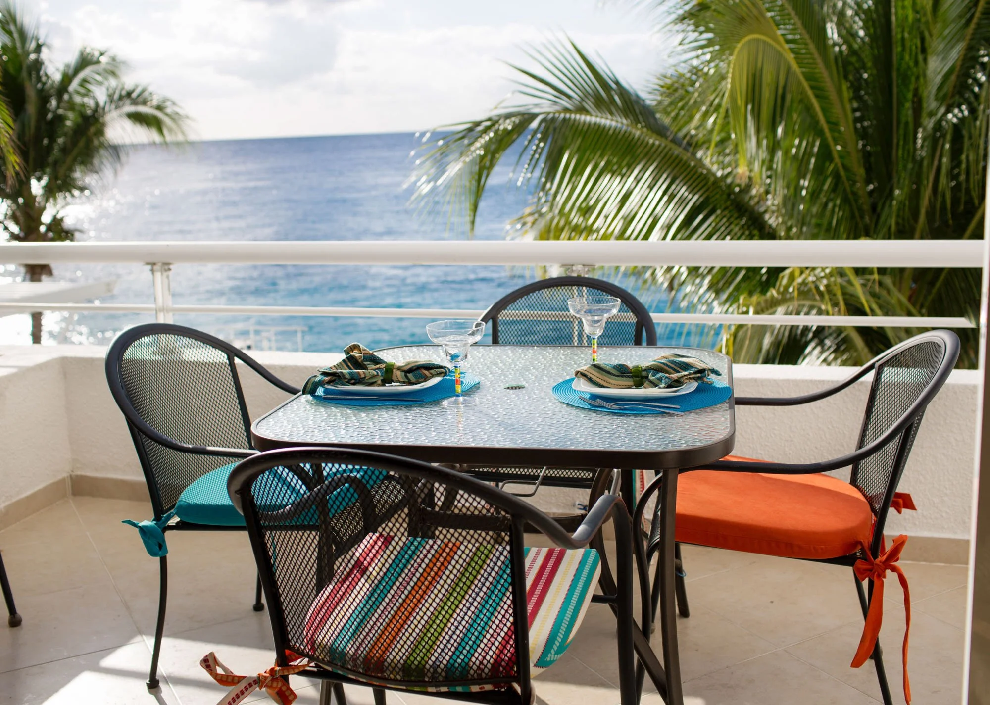 Outdoor dining table set for four with plates, utensils, and glasses on a balcony overlooking the ocean, with palm trees in the background.