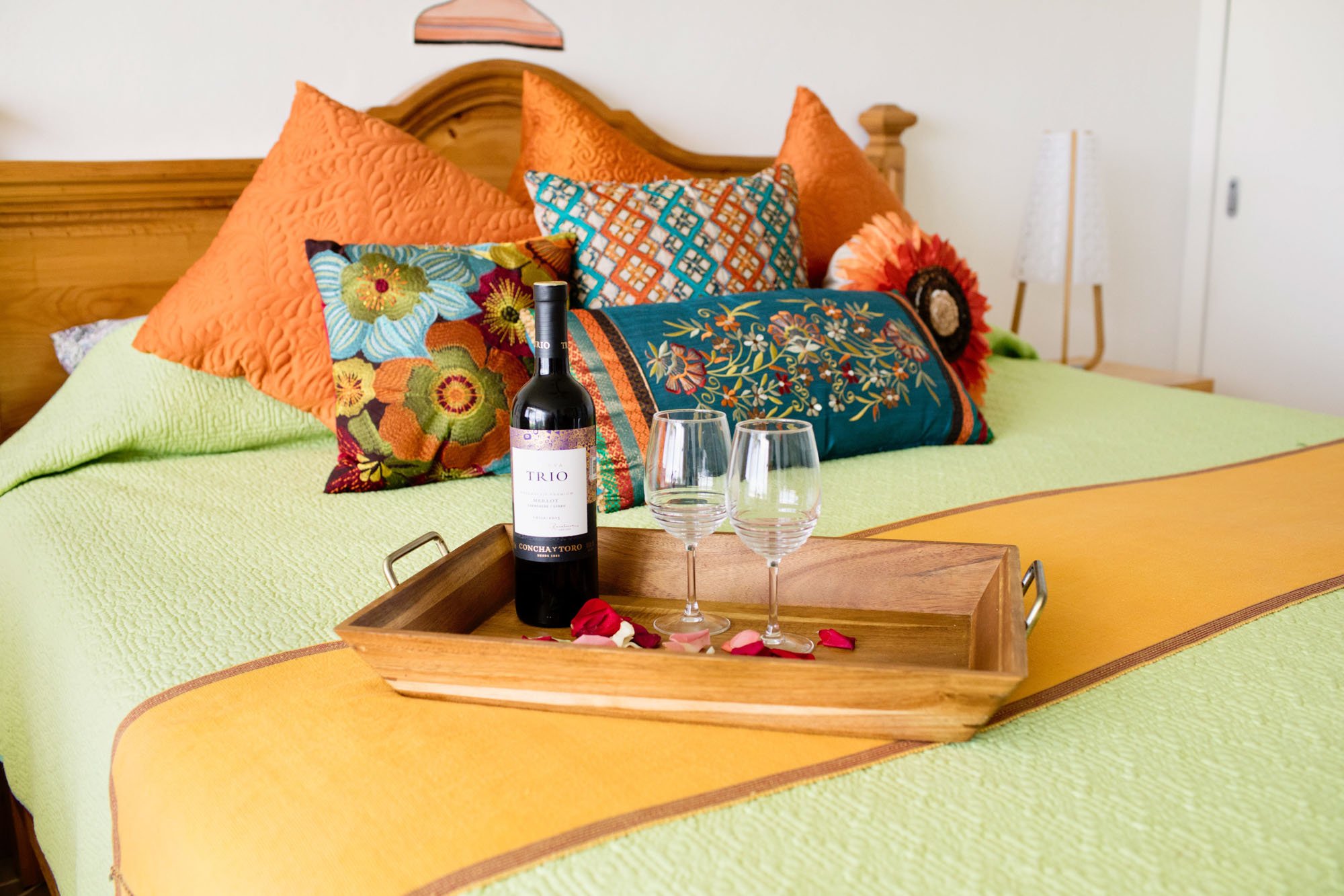 A wooden tray on a bed with a bottle of red wine, two empty wine glasses, and scattered rose petals, with colorful pillows and bedspread in the background.