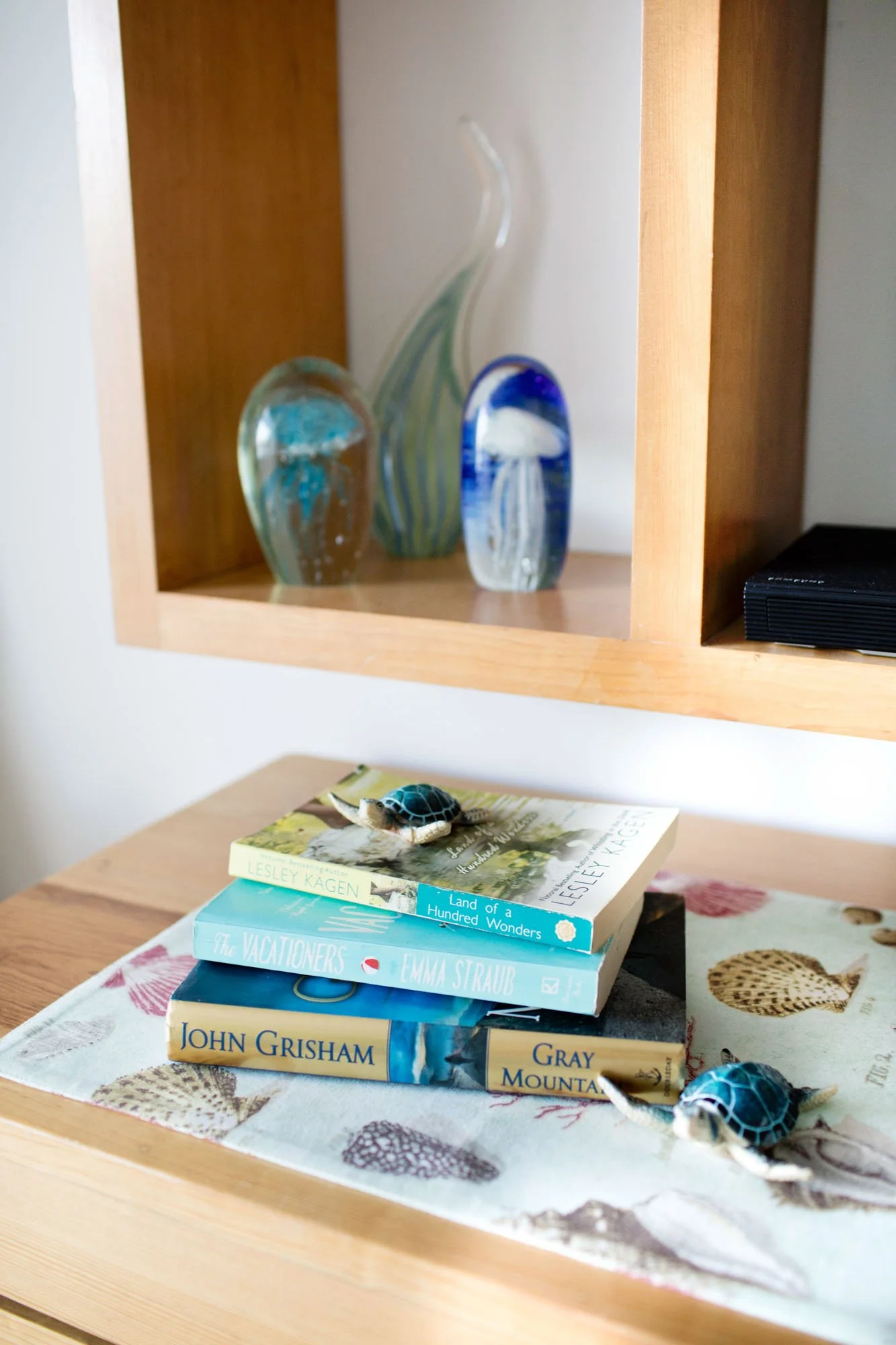 Decorative glass art pieces and a stack of books with toy turtles on top and beside it on a wooden table with a shell-themed tablecloth.