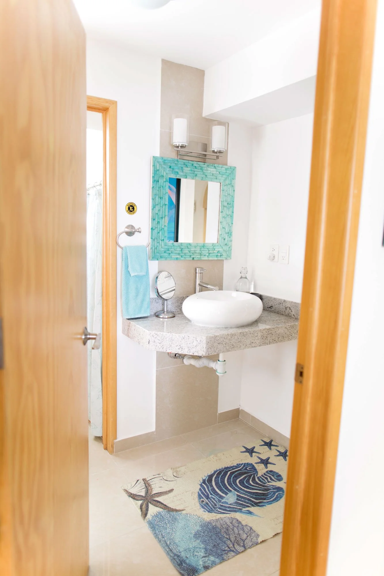 Bathroom with a circular vessel sink, small mirror, blue towel, turquoise framed mirror, wall-mounted light fixtures, ocean-themed rug, and wooden door frame.