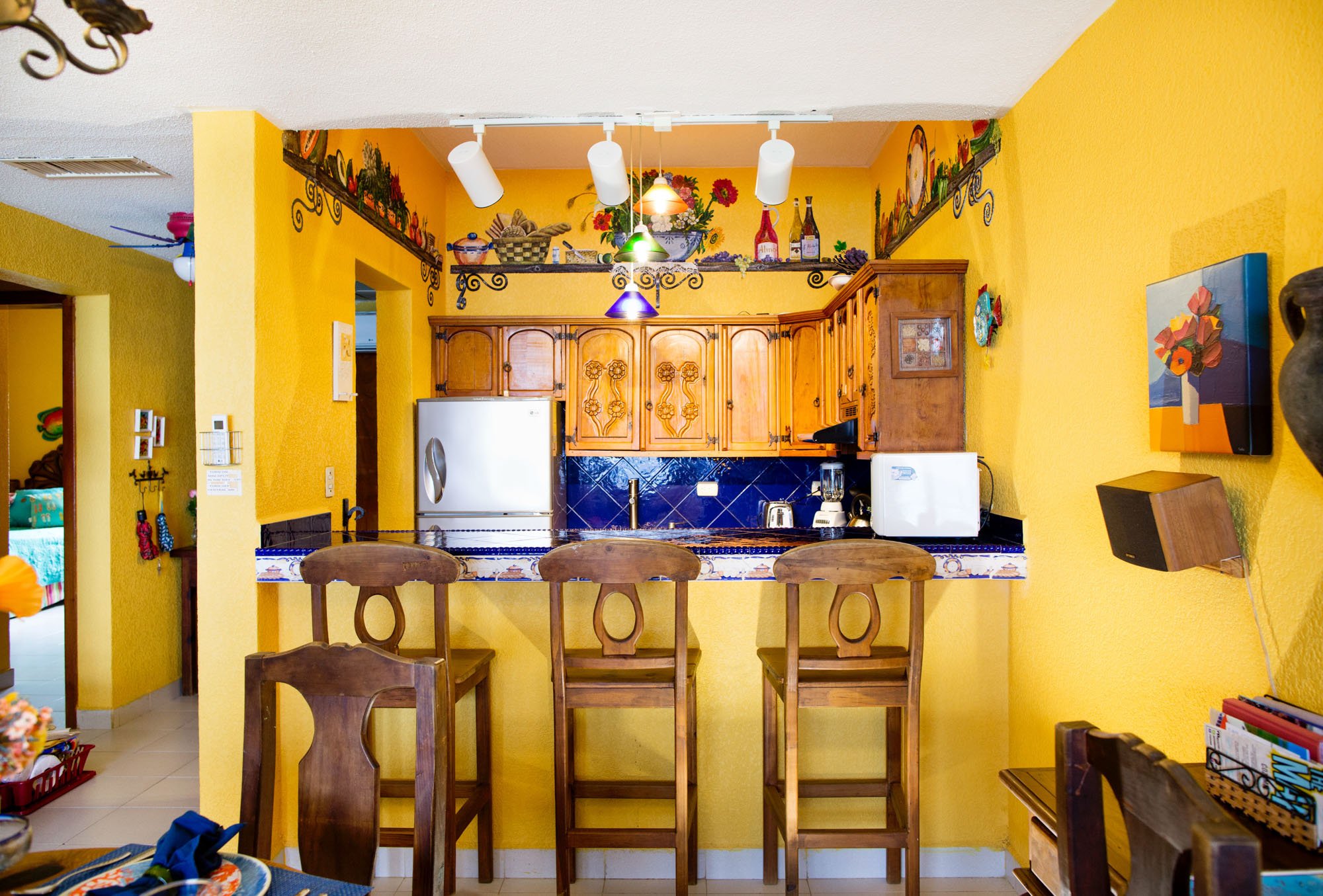 Cozumel vacation rental kitchen with yellow walls, wooden cabinets, a blue tiled backsplash, and a breakfast bar with four wooden chairs.