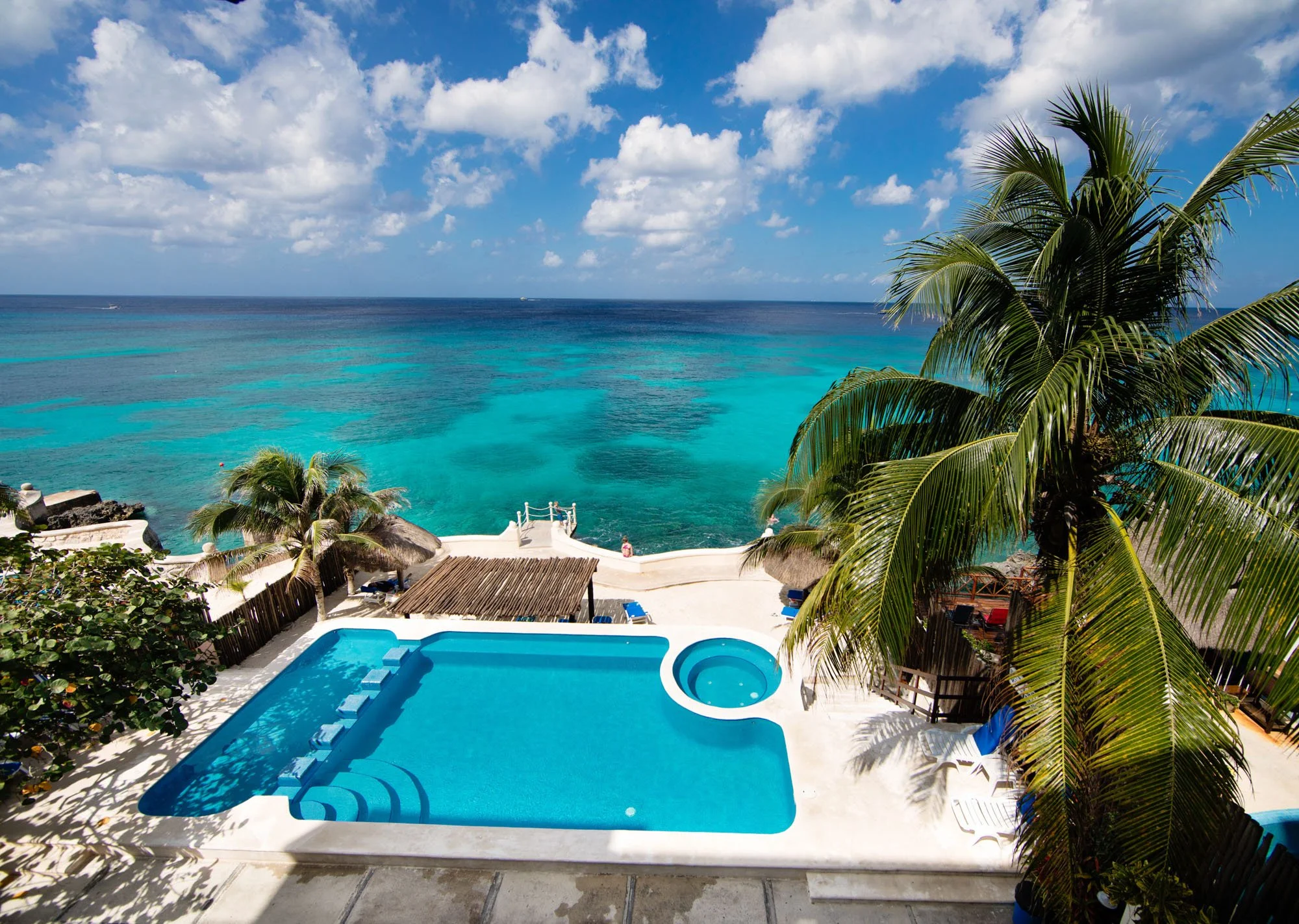 Cozumel vacation rental ocean view with turquoise waters, blue sky with scattered clouds, a swimming pool with a hot tub, and tropical palm trees surrounding the pool area.