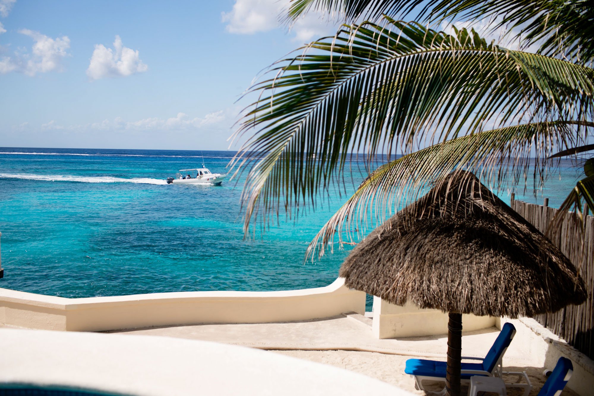 View of a tropical beach with a thatched hut, lounge chairs, palm trees, and a boat on turquoise water with a blue sky.