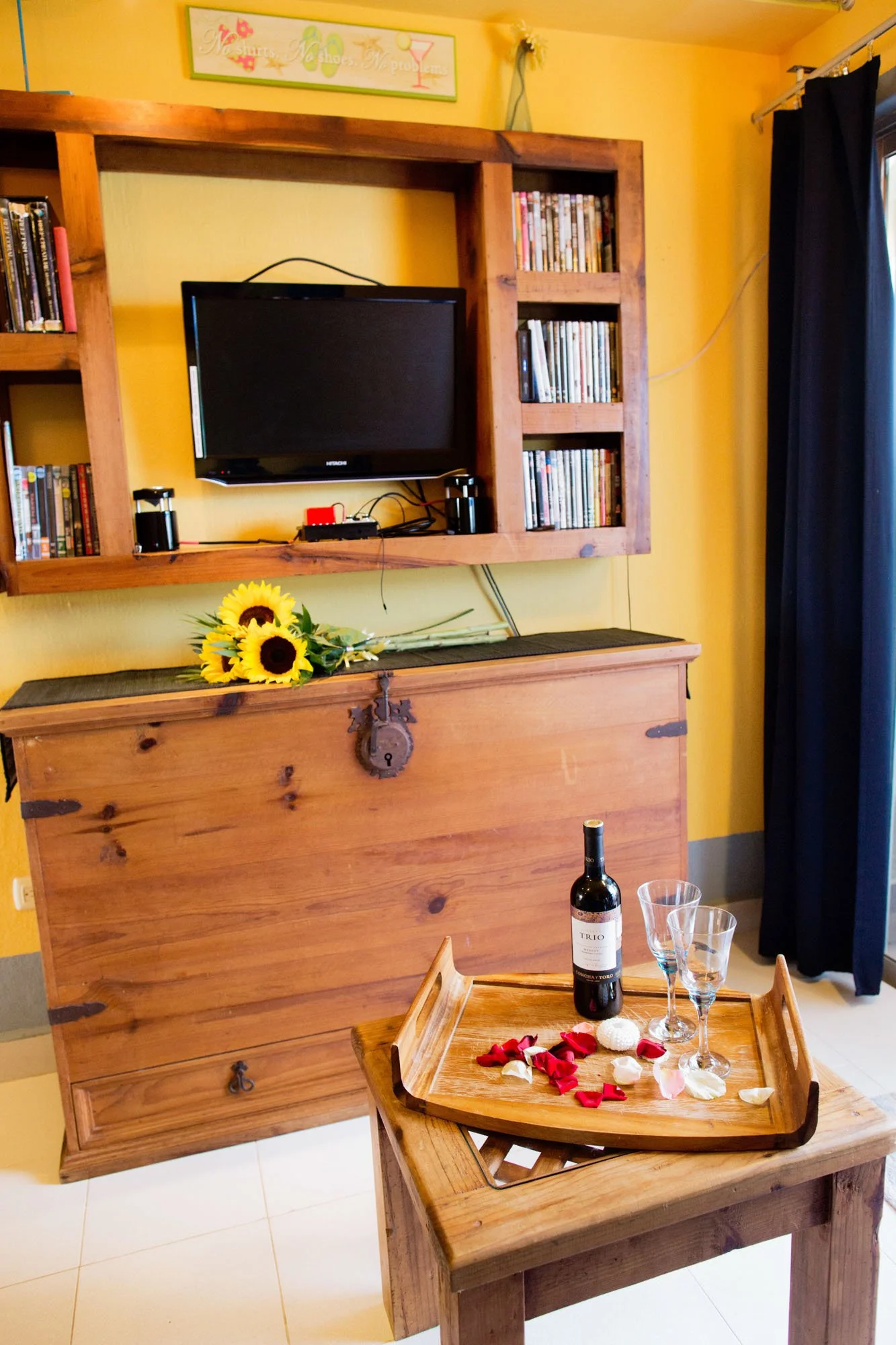 A cozy living room corner with a yellow wall, a rustic wooden cabinet with a lock, a small flat-screen TV, a bouquet of sunflowers on the cabinet, and a wooden tray on a small table with a bottle of red wine, two empty wine glasses, and rose petals.