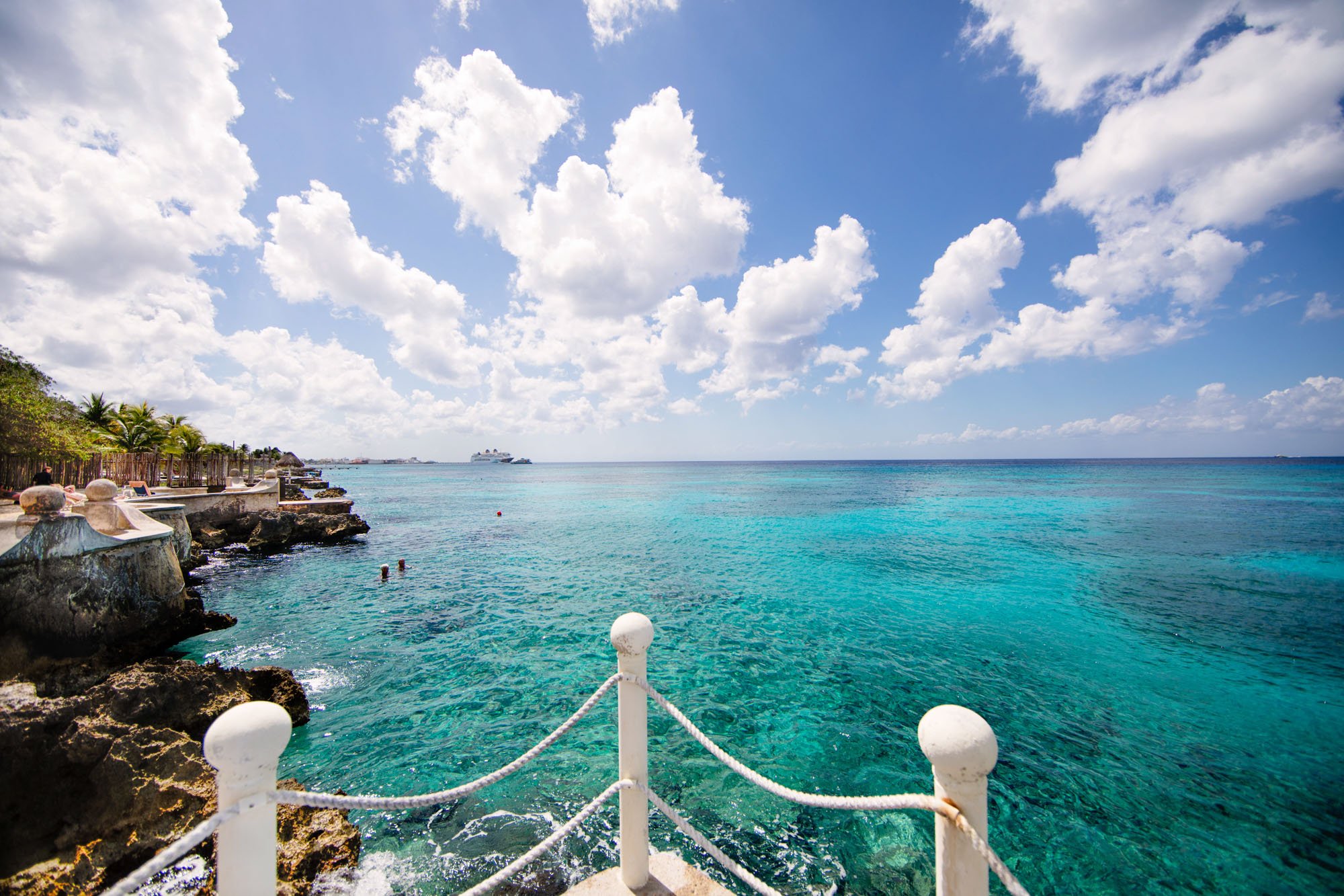 Cozumel vacation rental seaside view with clear blue water, rocky shoreline, palm trees, and ships in the distance under a partly cloudy sky.
