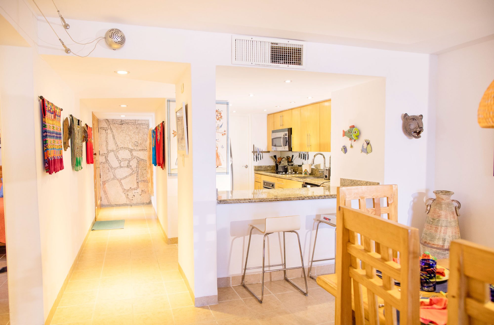 Kitchen and dining area with wooden chairs, art decorations on white walls, and a hallway with colorful textiles hanging on the wall at a Cozumel vacation rental.