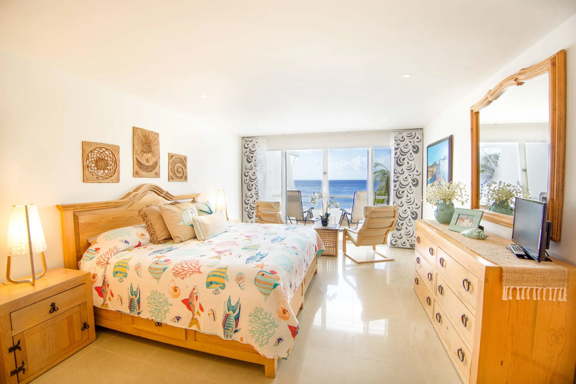 Bright bedroom with ocean view, wooden furniture, bed with colorful sea life themed bedding, and seating area by sliding glass doors