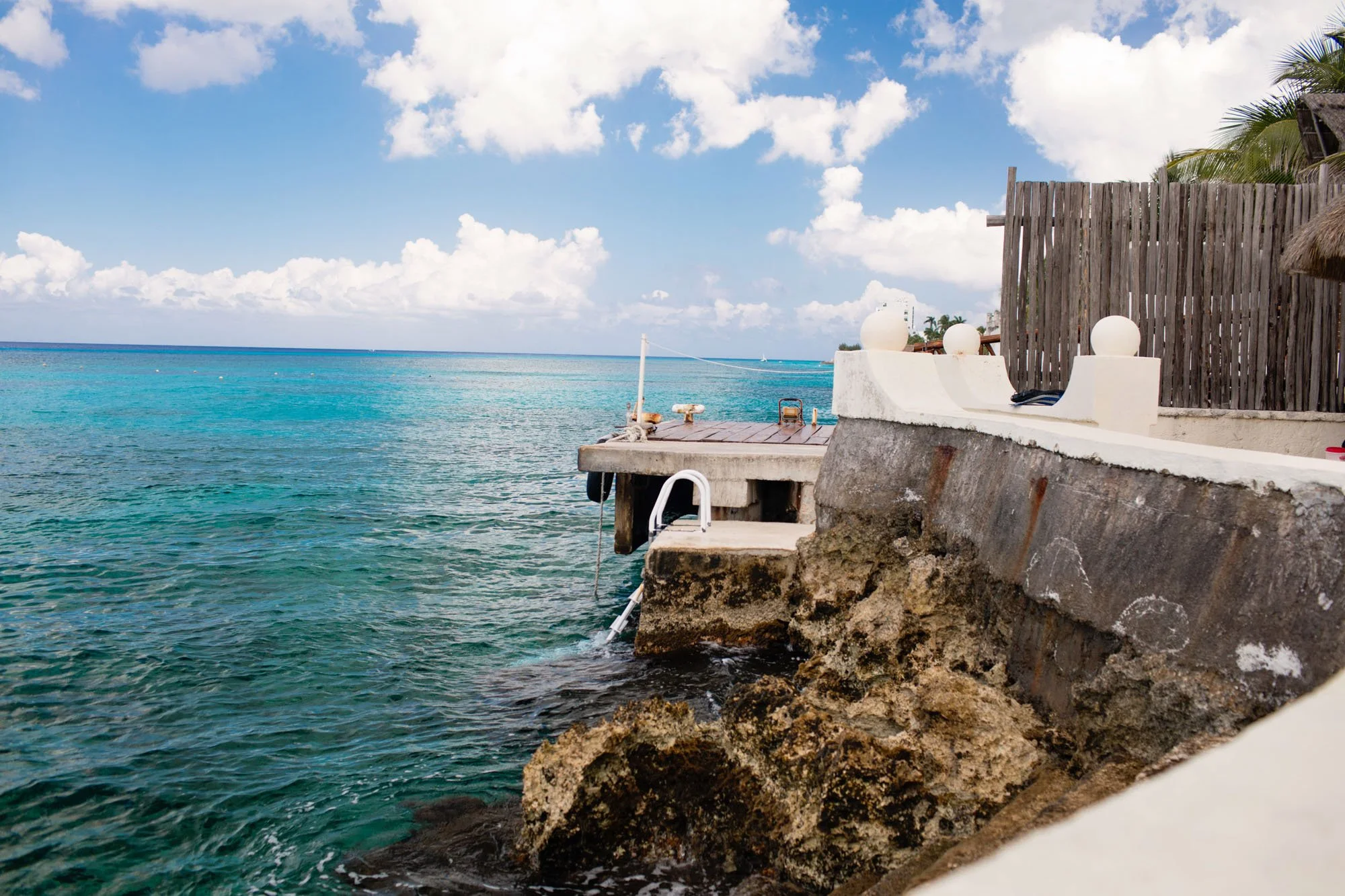 Photo of a tropical beach with turquoise waters, a concrete dock, and a wooden fence with large white spherical decorations on a sunny day with a blue sky and scattered clouds.