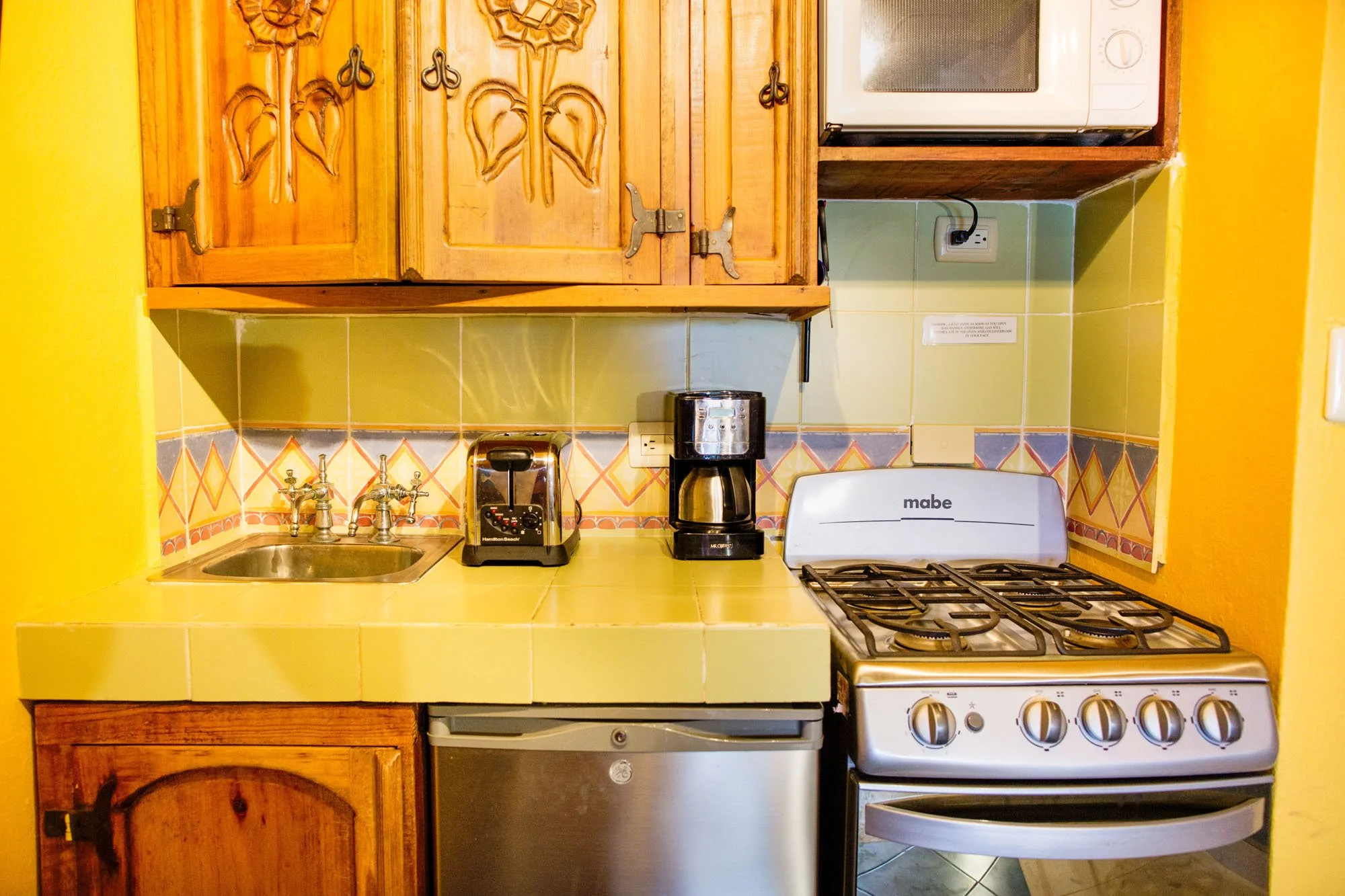 Small kitchen with wooden cabinets, yellow tiled countertop, and green tiled backsplash. Contains a microwave, coffee maker, toaster, and gas stove. Yellow walls.