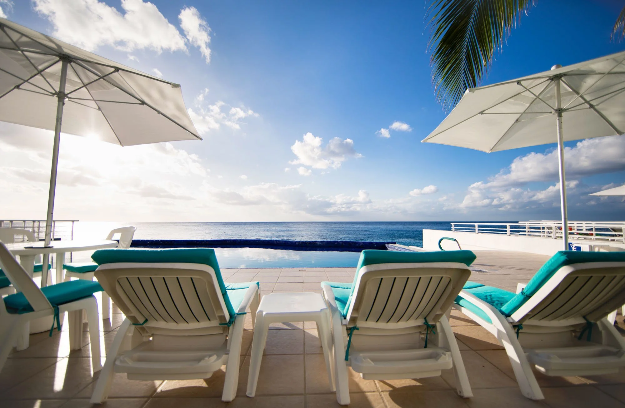 Poolside chairs with turquoise cushions and white umbrellas overlooking the ocean under a partly cloudy sky