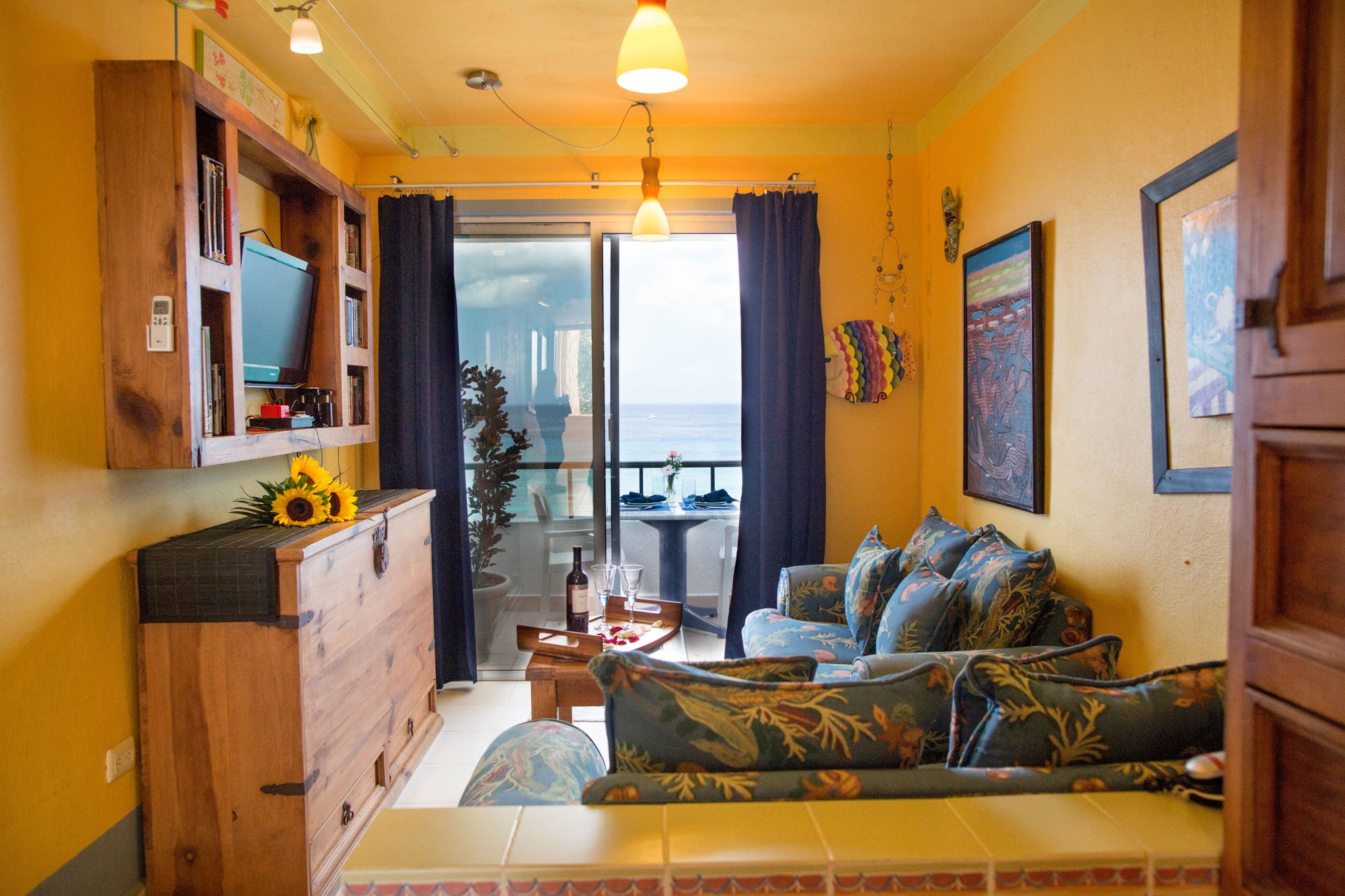 Living room with yellow walls, dark blue curtains, floral-patterned sofa, wall art, TV on a wooden shelf, and sliding glass door leading to a balcony with ocean view at a Cozumel vacation rental.