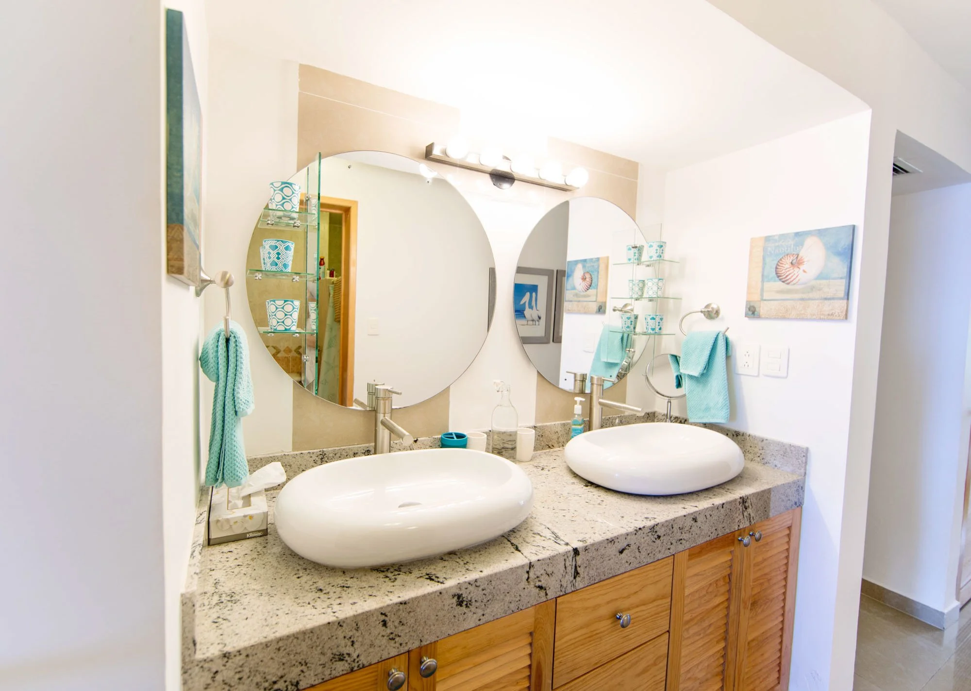 Bathroom with two round vessel sinks and large mirrors above, decorated with beach-themed paintings and turquoise towels.