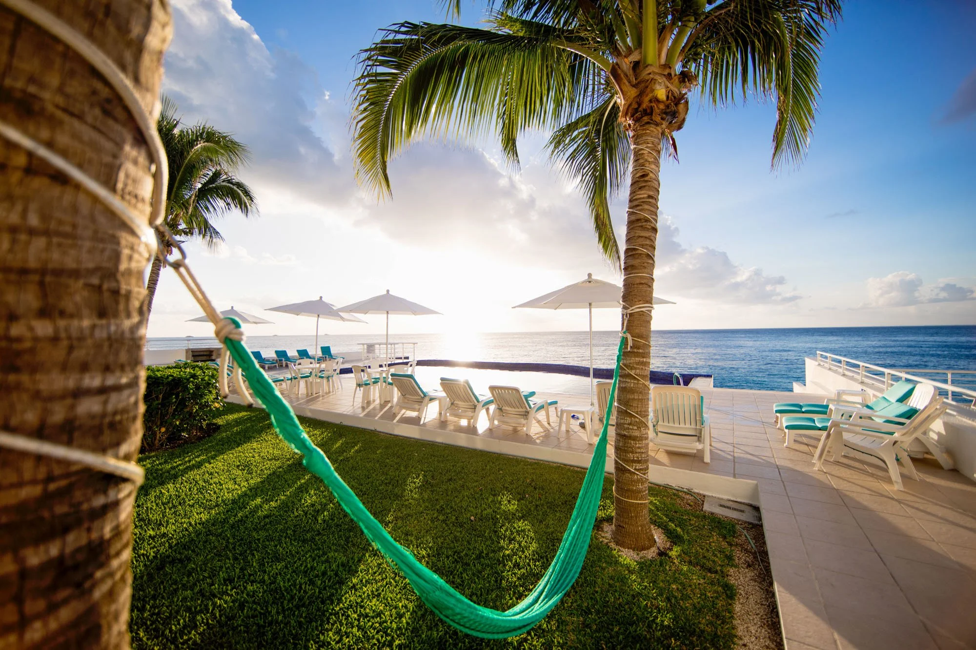 Sunset view overlooking the ocean with a poolside area, lounge chairs, umbrellas, and palm trees, with a hammock in the foreground at a Cozumel vacation rental.