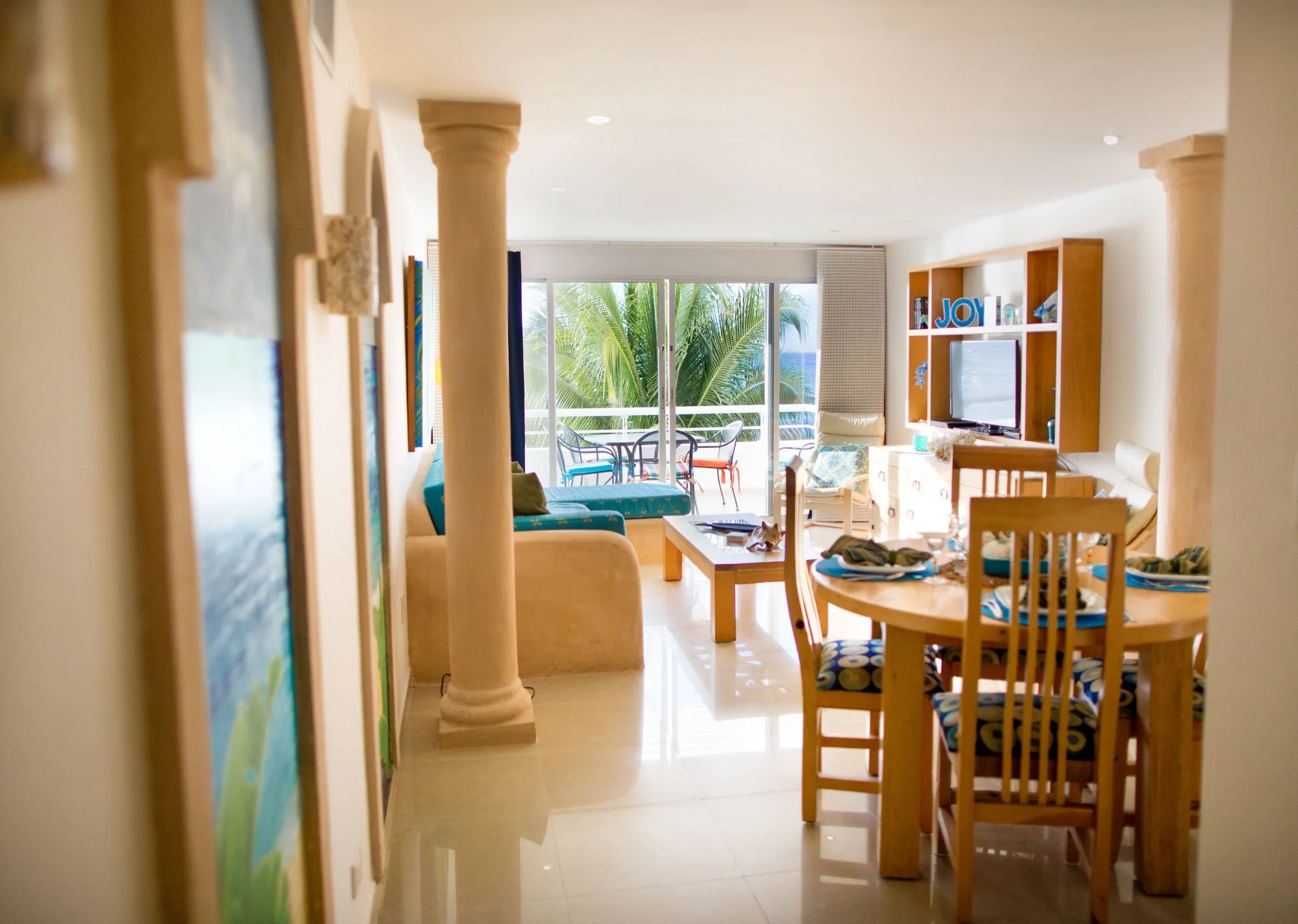 Living room and dining area with large sliding glass doors leading to a balcony with outdoor furniture and palm trees, furnished with a dining table, chairs, a sitting sofa, TV, and decorative items.