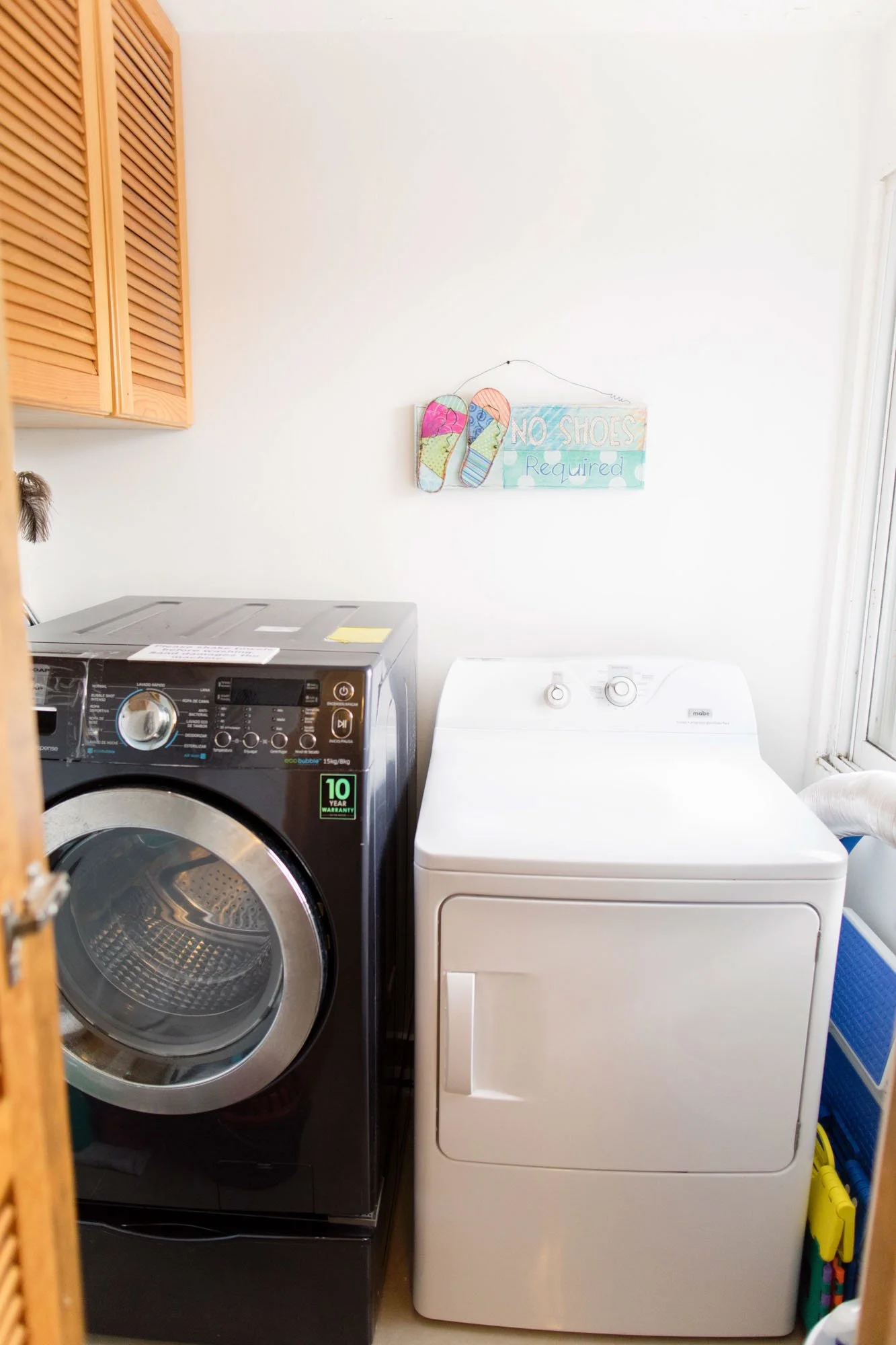 A laundry room with a black front-loading washing machine and a white top-loading dryer side by side, with a sign above that says "No Shoes Required" and a pair of colorful flip-flops hanging on the wall.