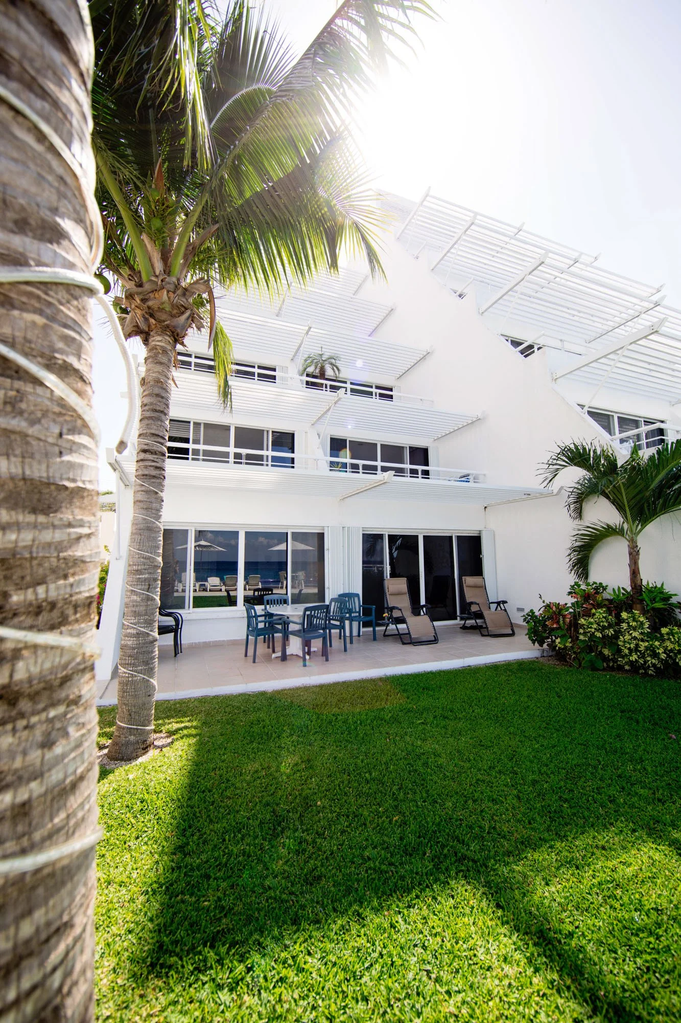 Cozumel vacation rental with outdoor patio furniture, lounge chairs, palm trees, manicured lawn, and glass sliding doors, under bright sunlight.
