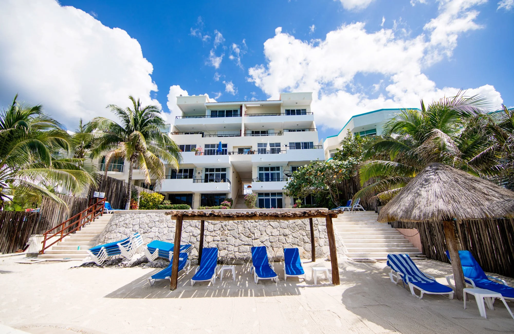 Beachside Cozumel vacation rental with palm trees, blue sky with clouds, beach chairs, and thatched umbrellas.