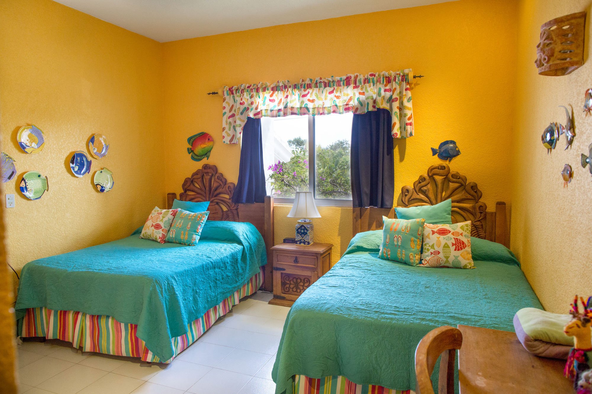 A bedroom with yellow walls, two wooden carved headboards, and two beds with teal bedspreads, patterned pillows, and multicolored striped bed skirts. There's a window with blue curtains and a colorful valance, a wooden nightstand with a lamp, and dec