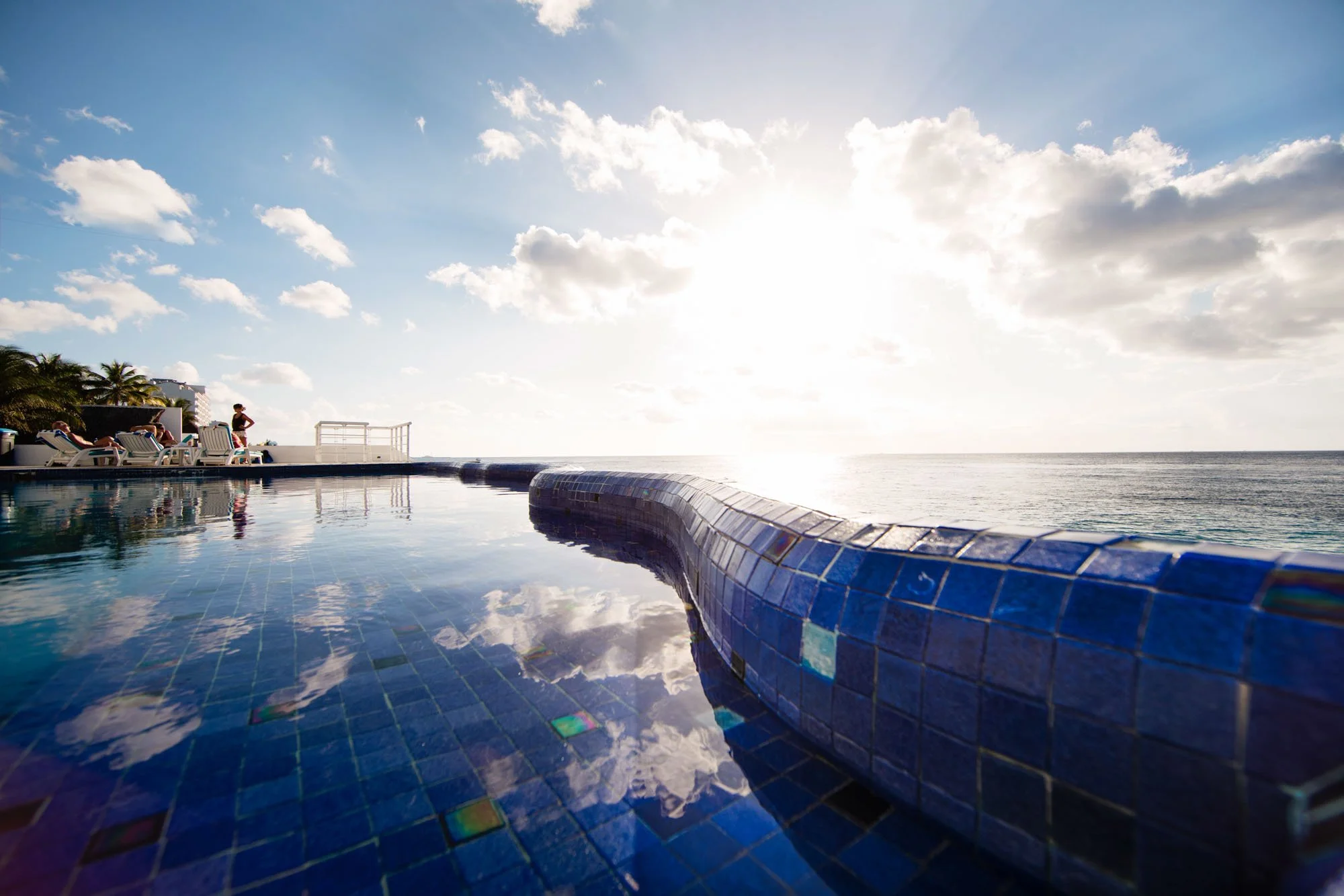 Swimming pool with blue mosaic tiles reflecting the sky and clouds, overlooking the ocean with lounge chairs and people relaxing in the background, under a bright sun and partly cloudy sky.
