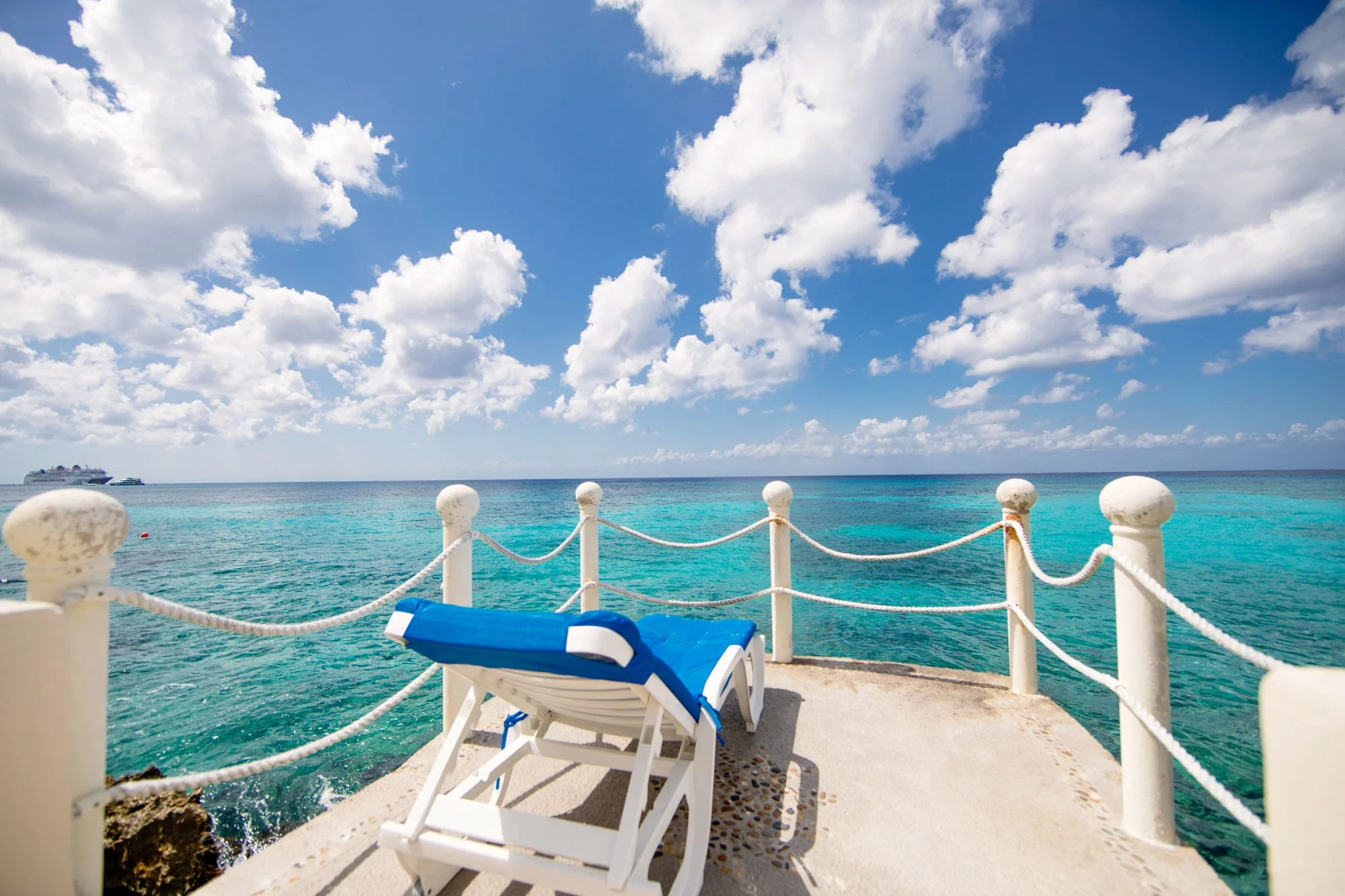 A white lounge chair with a blue cushion on a concrete pier, overlooking turquoise ocean water with a cruise ship in the distance, under a partly cloudy blue sky.