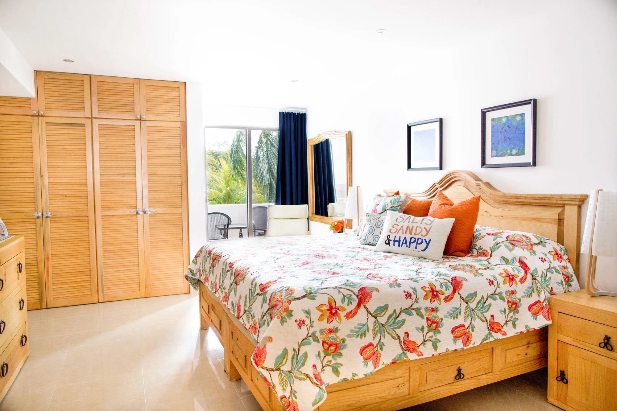 Bright hotel bedroom with a large bed covered in a floral quilt and decorative pillows, wooden furniture, a sliding glass door leading to a balcony with outdoor chairs, a large mirror, and framed artwork on white walls.