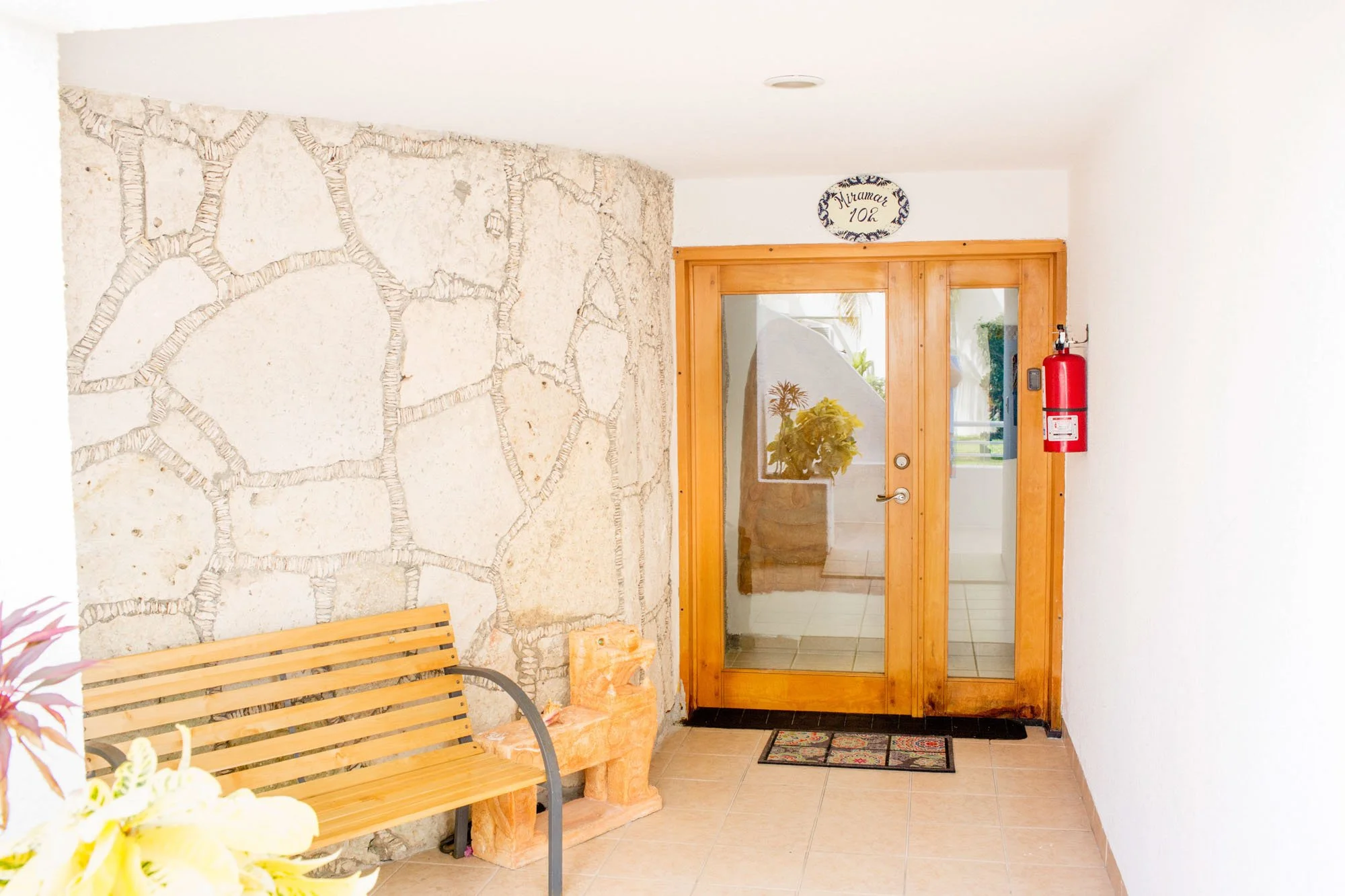 Cozumel vacation rental entrance with wooden door, stone wall, fire extinguisher, and a bench.