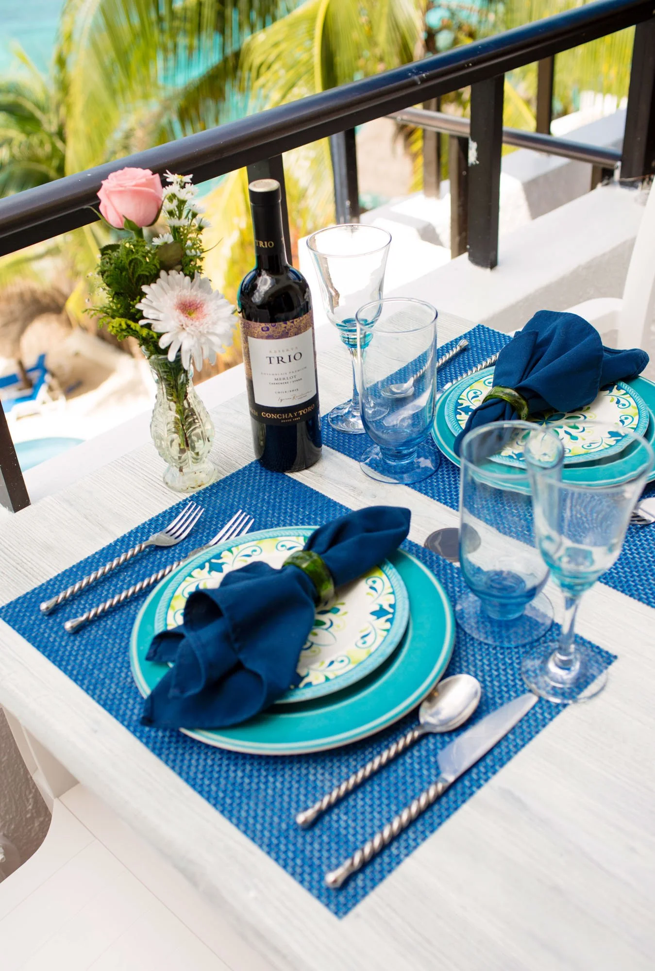 Dining table set for four with blue placemats, patterned plates, and navy blue cloth napkins with green napkin rings. A bottle of red wine and five glasses are on the table. There is a small flower arrangement in a glass vase, with pink and white flo