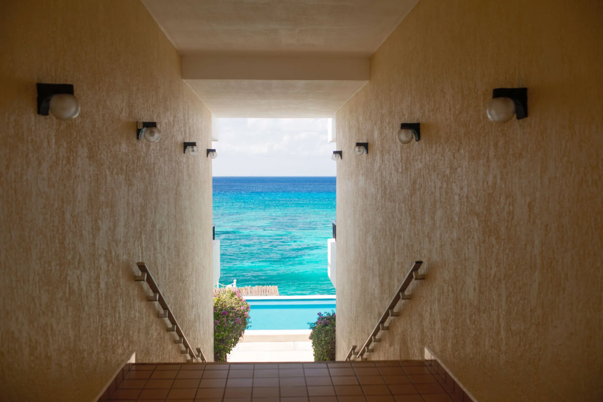 View from a corridor overlooking the ocean, with a pool area and colorful flowers in the foreground.