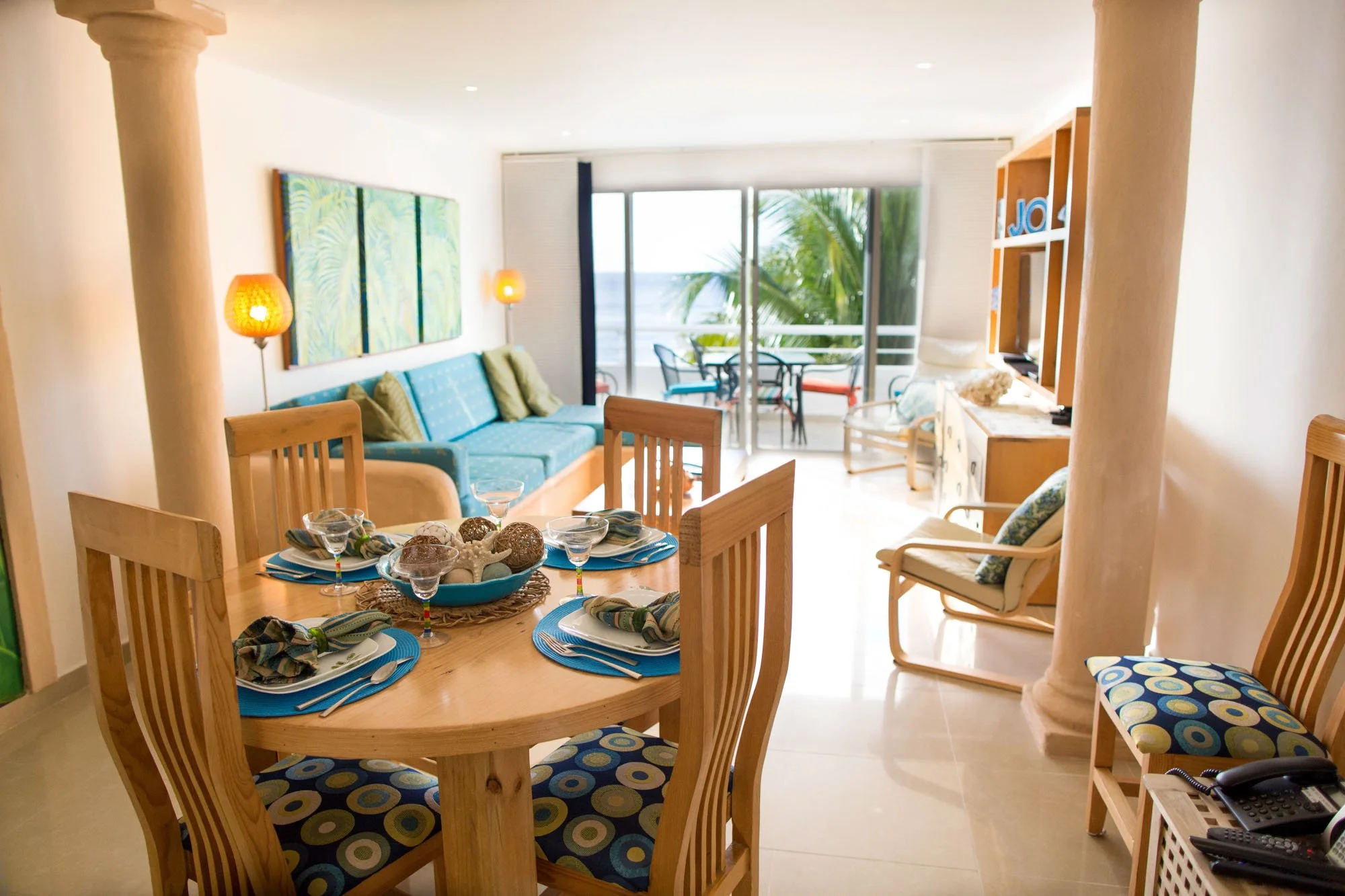 Living room with a round dining table set with plates, napkins, and bowls, a blue sofa with throw pillows, a rocking chair, a TV on a cabinet, and a balcony with outdoor furniture and a view of palm trees.