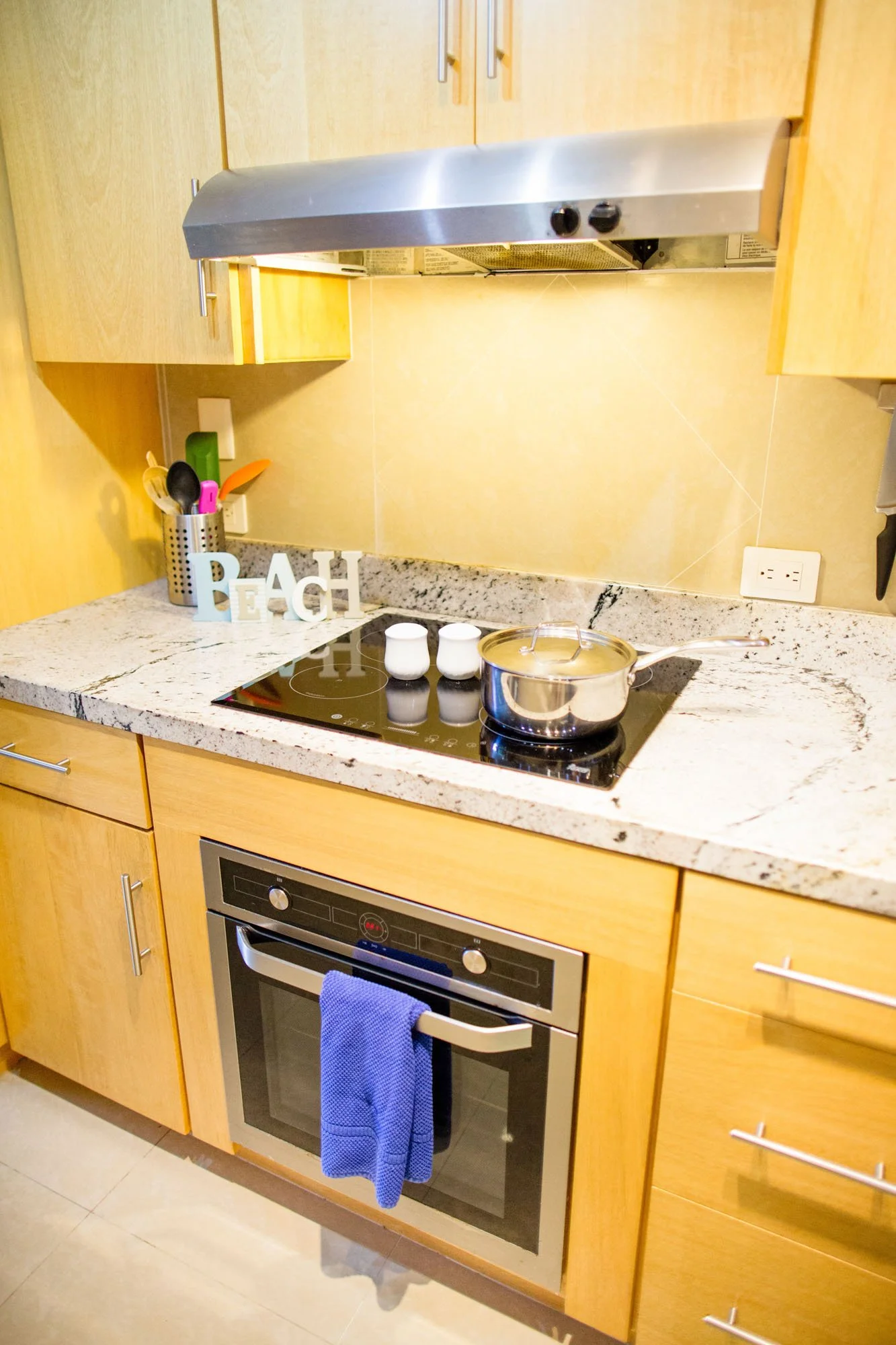 Kitchen stove with a pot on the cooktop, kitchen utensils in a container, decorative "Beach" sign, wooden cabinets, built-in oven with a blue towel hanging, and beige tiled backsplash.