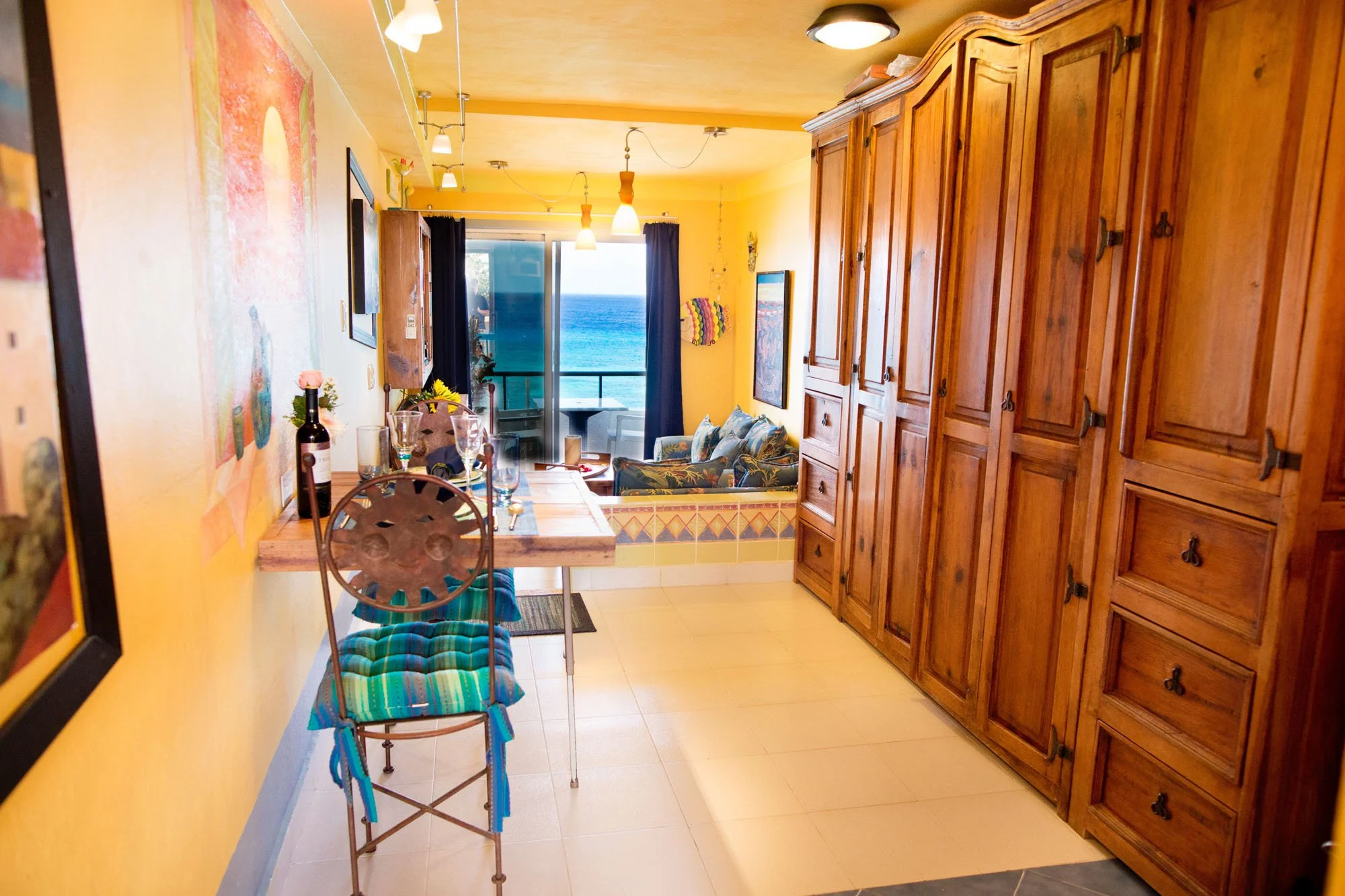 View of a brightly lit condo living area with a view of the ocean, featuring wooden cabinets, a seating area with a view, and a dining table set for a meal at a Cozumel vacation rental.